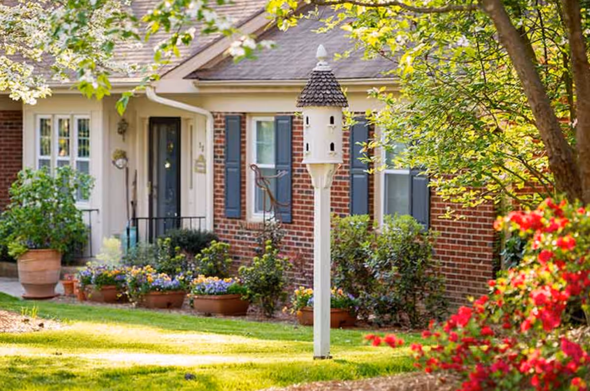 Front exterior of a brick building with a white decorative birdhouse post, potted flowers, shrubs, and an entry door.