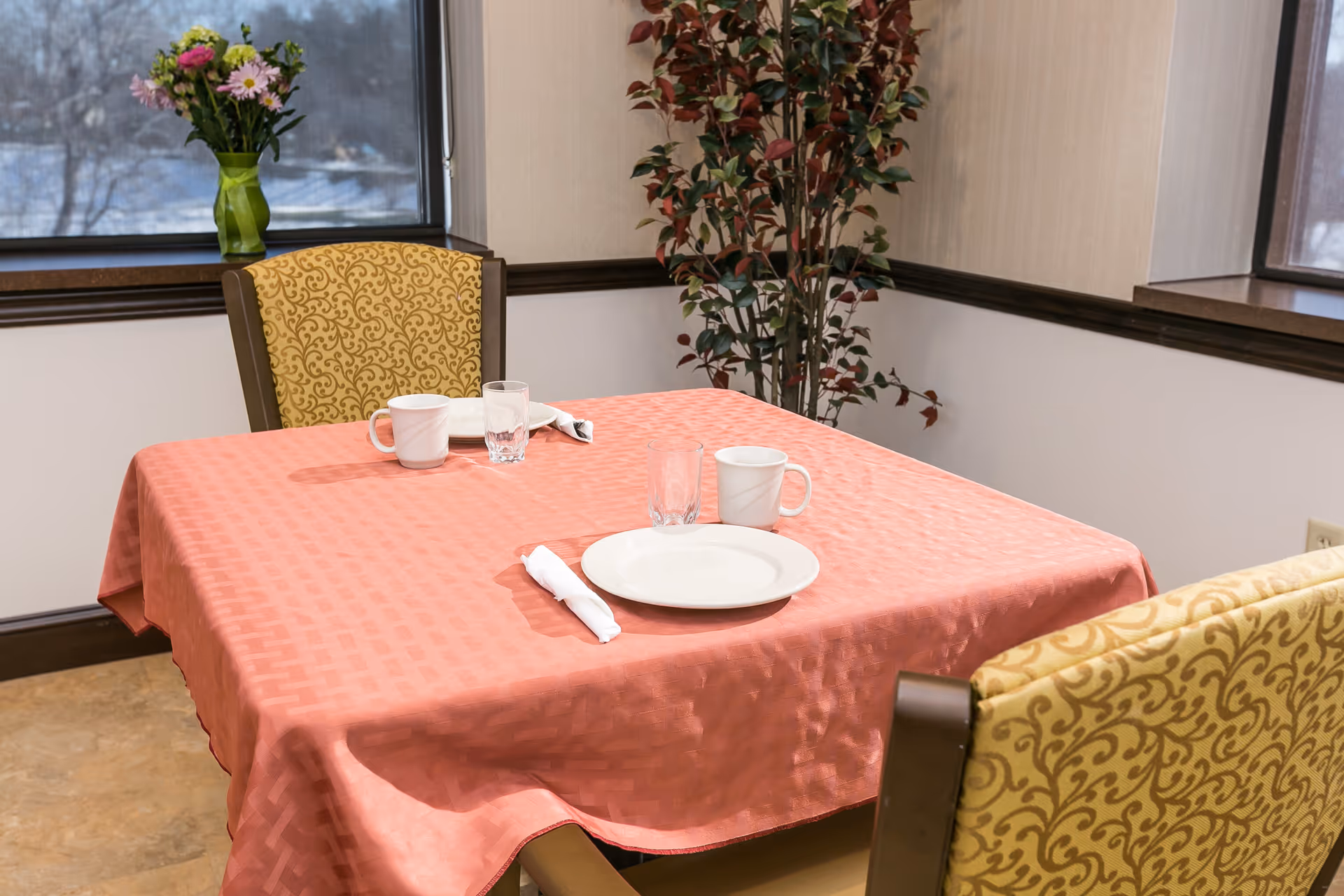 A small dining table covered with a salmon-colored tablecloth set for two with white plates, mugs, glasses, and rolled napkins. Two upholstered chairs with yellow patterned fabric are positioned at the table. A vase with flowers is on the windowsill, and a tall leafy plant is in the corner near the window.
