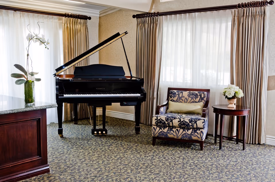 Sunlit lounge area with a black grand piano, patterned armchair, small side table with flowers, and large curtained windows.