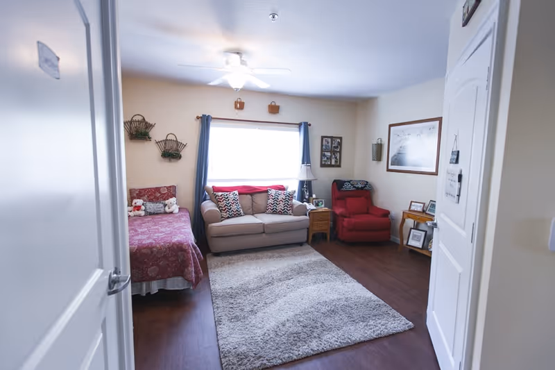 A cozy senior living room with a beige sofa adorned with patterned pillows, a red recliner chair, and a single bed with a red patterned bedspread and stuffed animals. The room has a large window with blue curtains, a ceiling fan with light, a soft gray area rug on wooden flooring, and various wall decorations including framed pictures and hanging baskets.