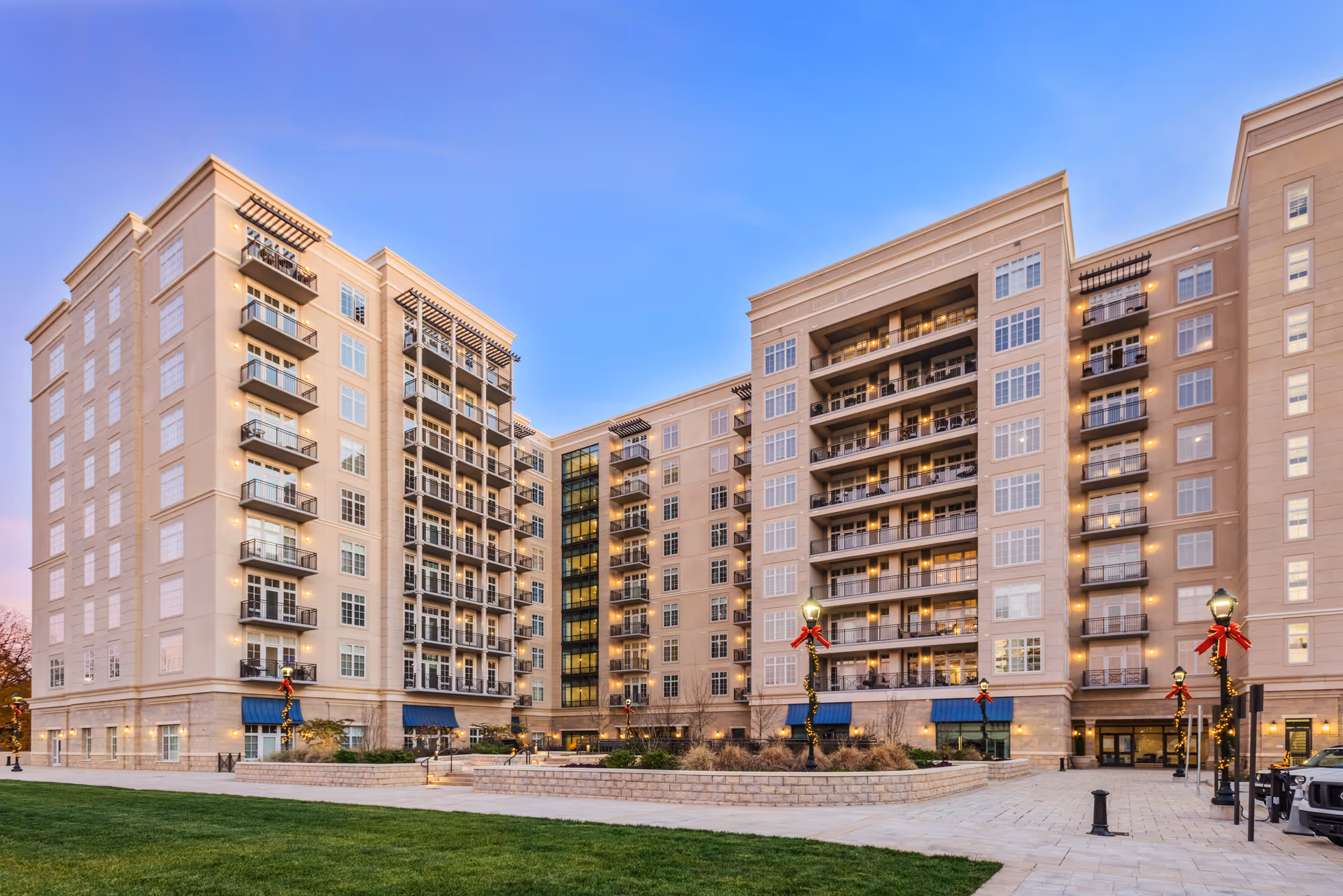 Large multi-story senior living building with balconies and lit windows at dusk, a paved courtyard and lamp posts decorated for the holidays.