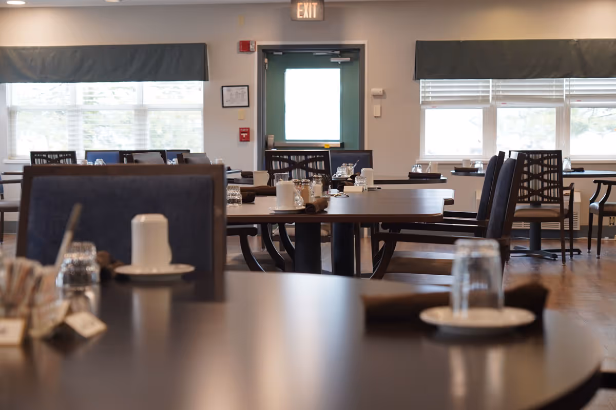 Interior view of a dining room with multiple tables and chairs arranged neatly. Tables are set with glasses, plates, and napkins. Large windows with blinds allow natural light to fill the room. An exit door is visible at the back of the room.