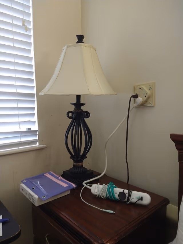A bedside table with a decorative lamp, a purple binder, and a power strip with cords plugged in. The table is next to a window with white blinds and a wall outlet with two plugs.