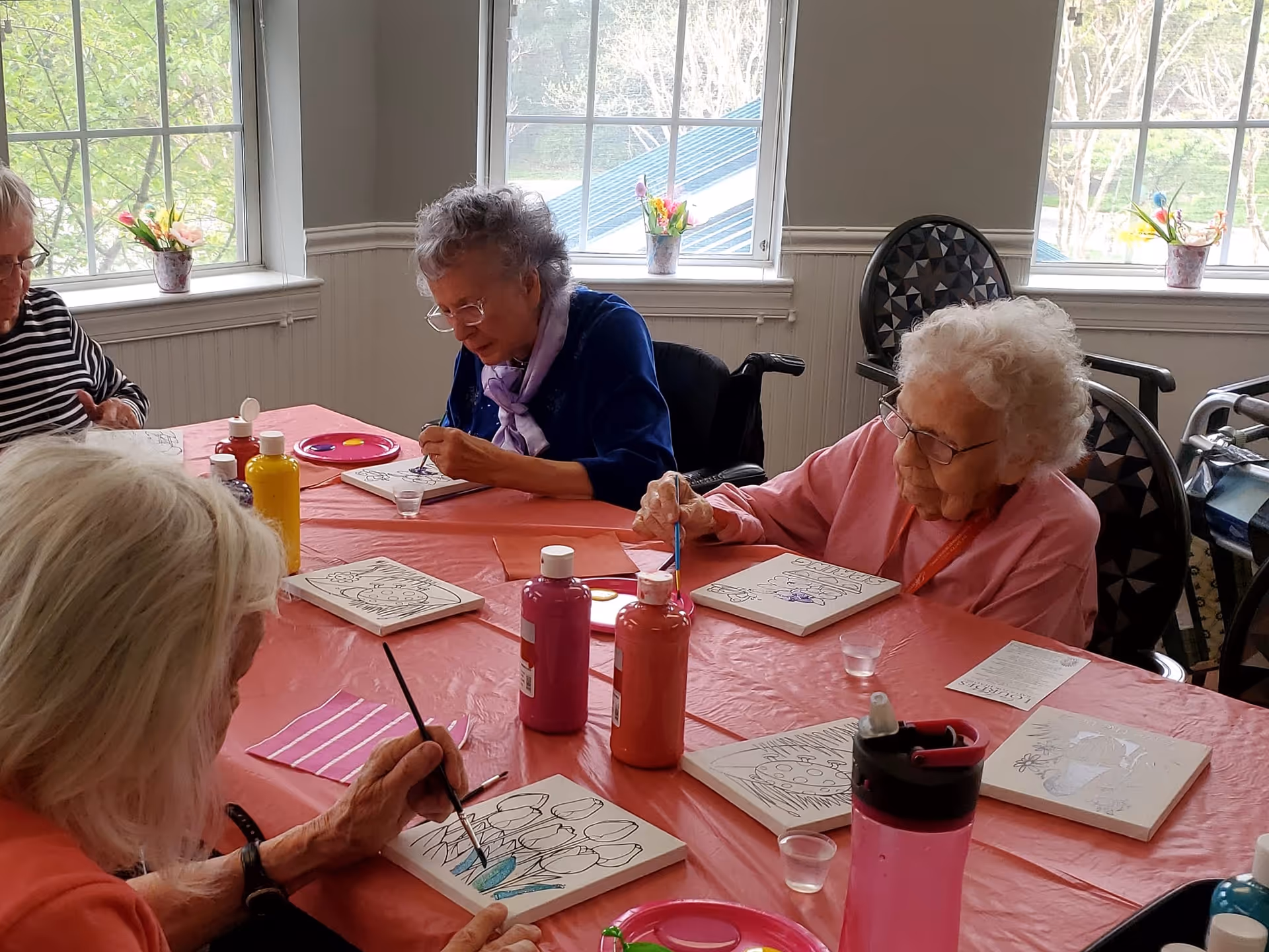 Four elderly women seated around a table covered with a pink tablecloth, engaged in painting on canvases. Various bottles of paint and small cups of water are on the table. The room has large windows with flower pots on the sills, letting in natural light.