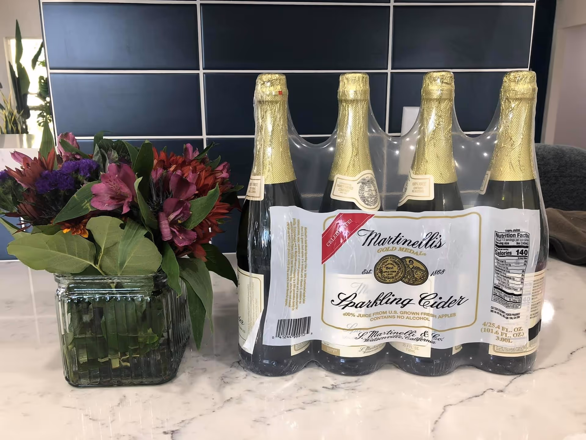 A clear glass vase with a bouquet of colorful flowers next to a six-pack of Martinelli's Gold Medal Sparkling Cider bottles on a marble countertop with a dark tiled backsplash in the background.