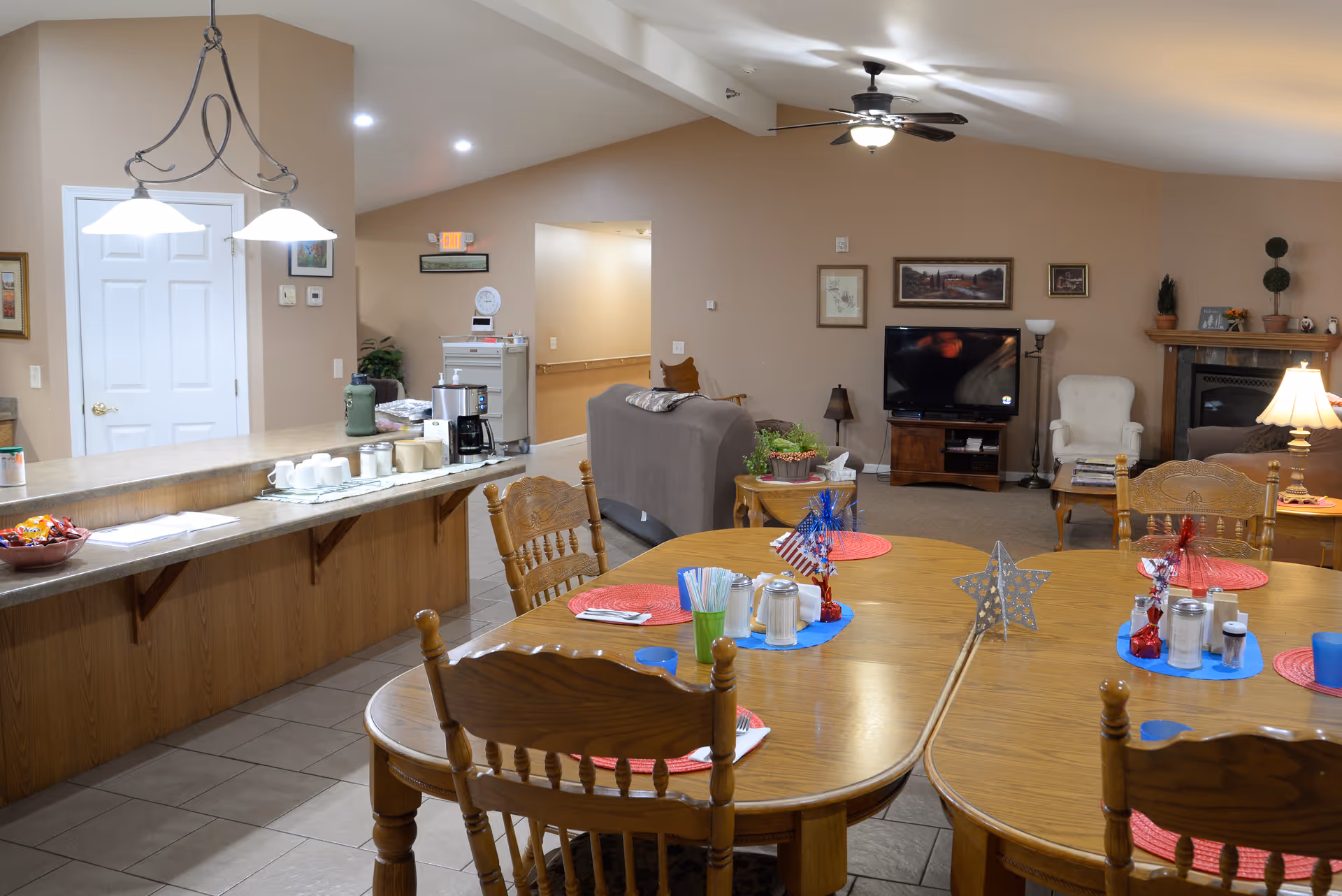Open dining and living area with wooden tables set with placemats and decorations, a counter with a coffee station, and seating and TV in the background.