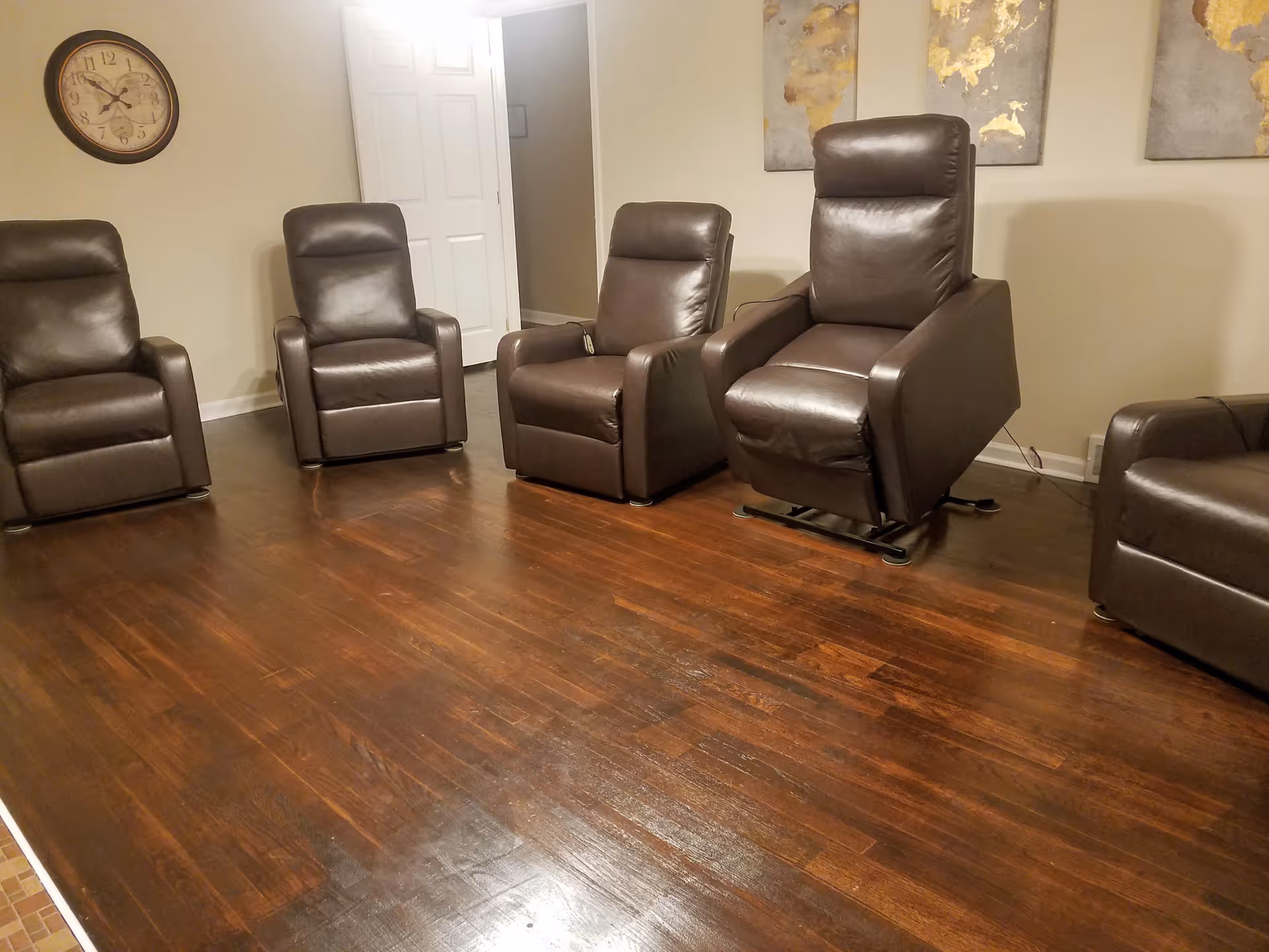 A room with five brown leather recliner chairs arranged in a semi-circle on a polished wooden floor. There is a wall clock on the left wall and three abstract gold and gray paintings on the right wall. A white door is partially open in the background.