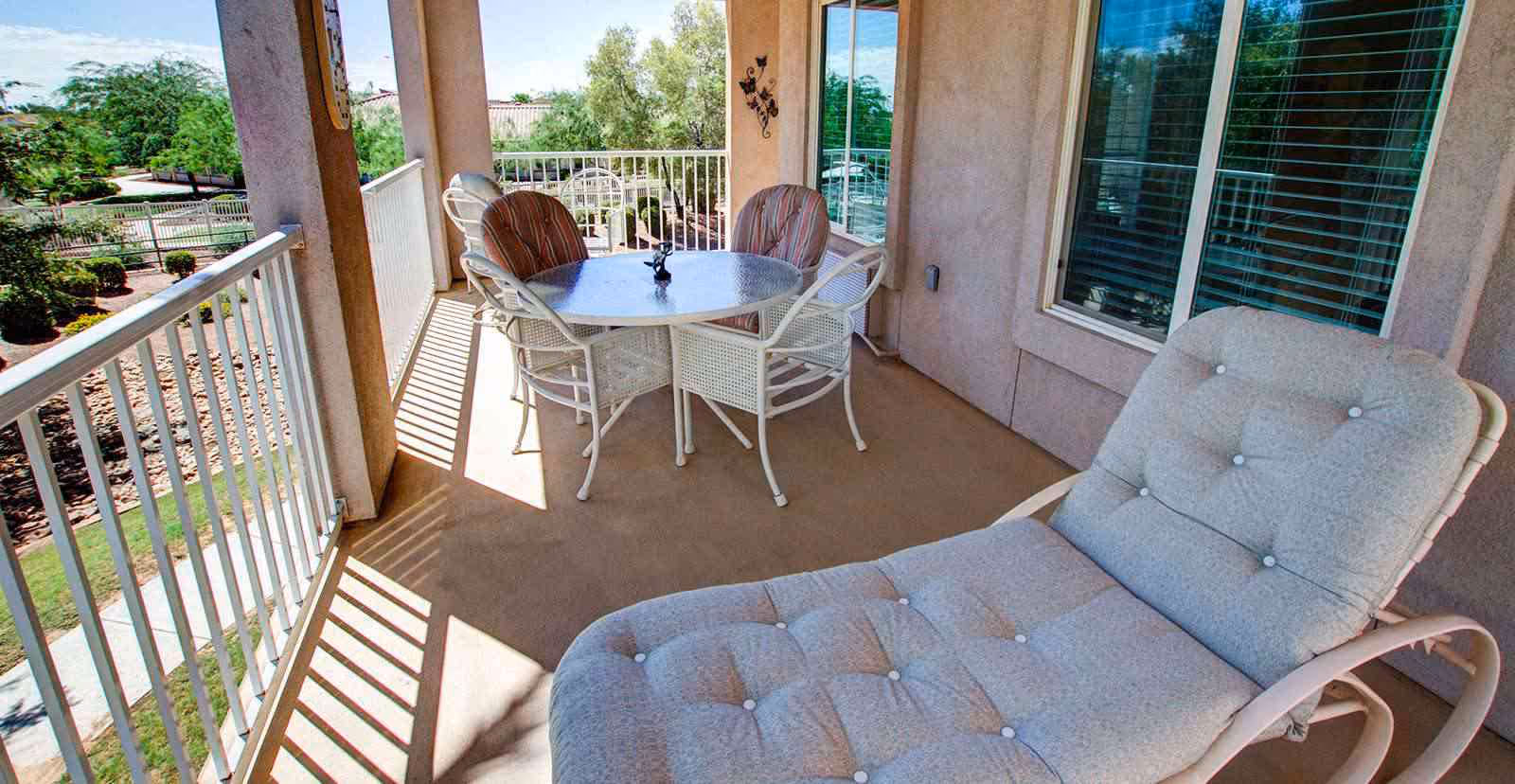 A covered outdoor balcony with a round glass table surrounded by four chairs, two of which have striped cushions. There is also a cushioned lounge chair in the foreground. The balcony has a white railing and overlooks a landscaped garden with trees and shrubs.