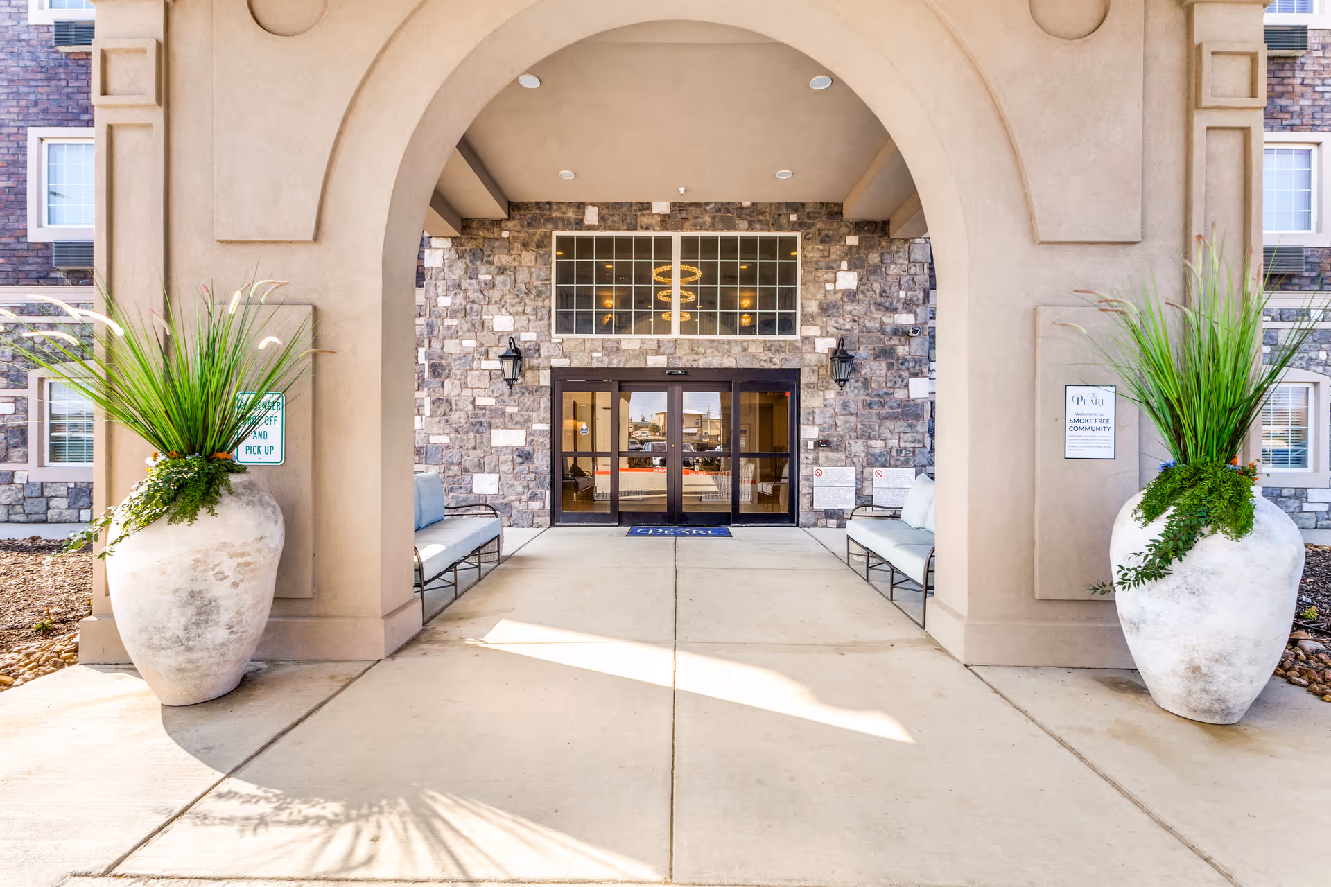 Entrance to The Pearl Senior Living at Lewisville featuring a stone facade with large glass doors under an archway. Two large white planters with green plants flank the entrance, and there are benches on either side of the doorway.