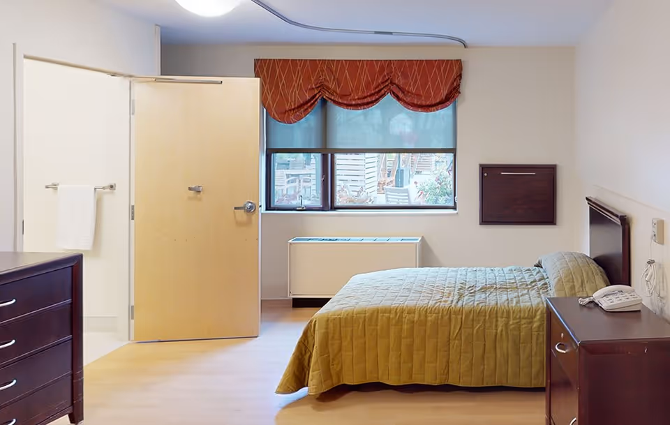 A simple bedroom in a senior living facility with a single bed covered in a green quilt, a wooden nightstand with a telephone, a wooden dresser, and a window with a red valance and blue roller shade. The door to an adjacent bathroom is open, showing a towel hanging inside.