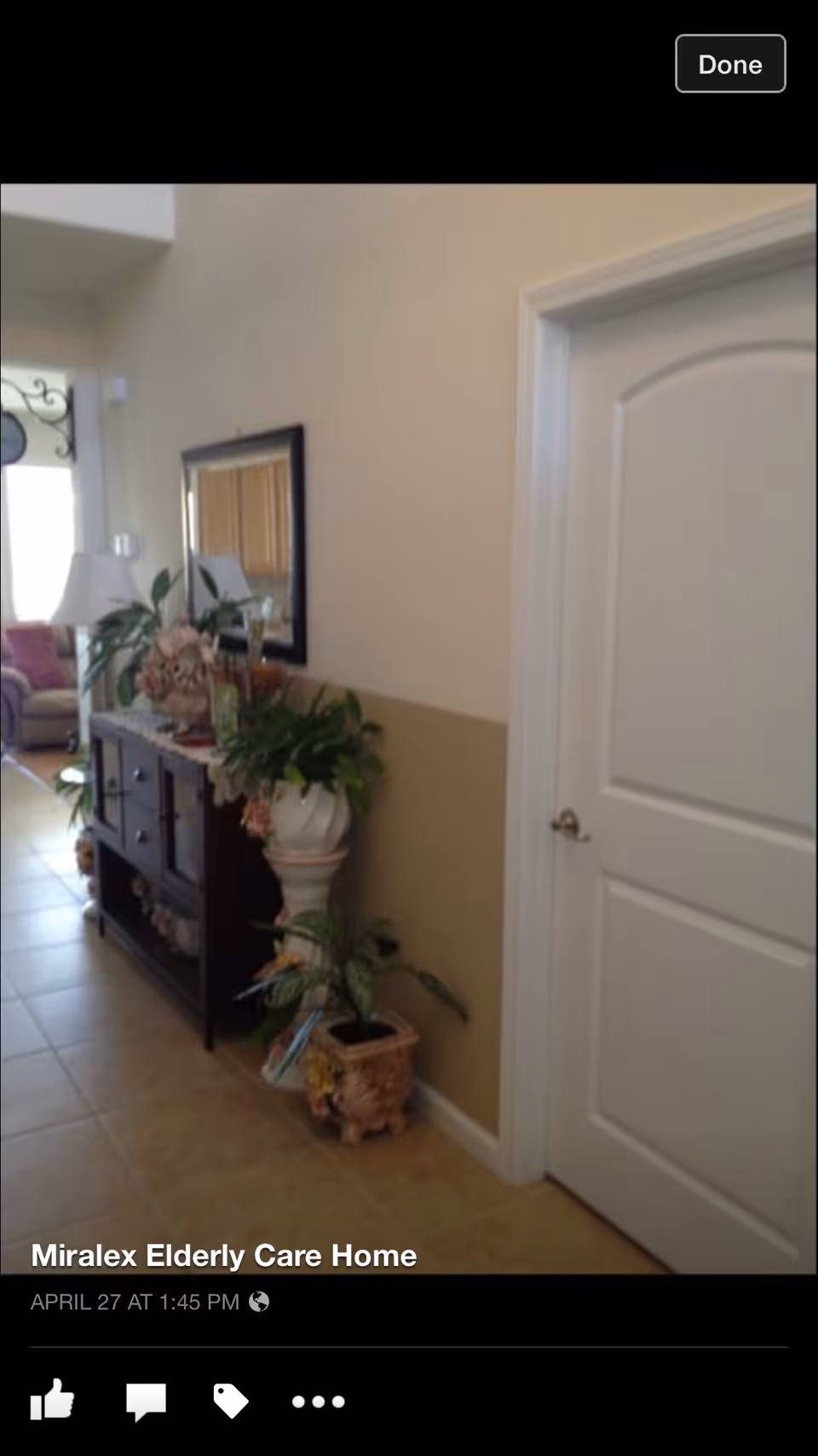 Interior hallway of Miralex Elderly Care Home with a white door on the right, a dark wooden console table with decorative items and plants on the left, a large mirror above the table, and a glimpse of a living area with a sofa and lamp in the background.