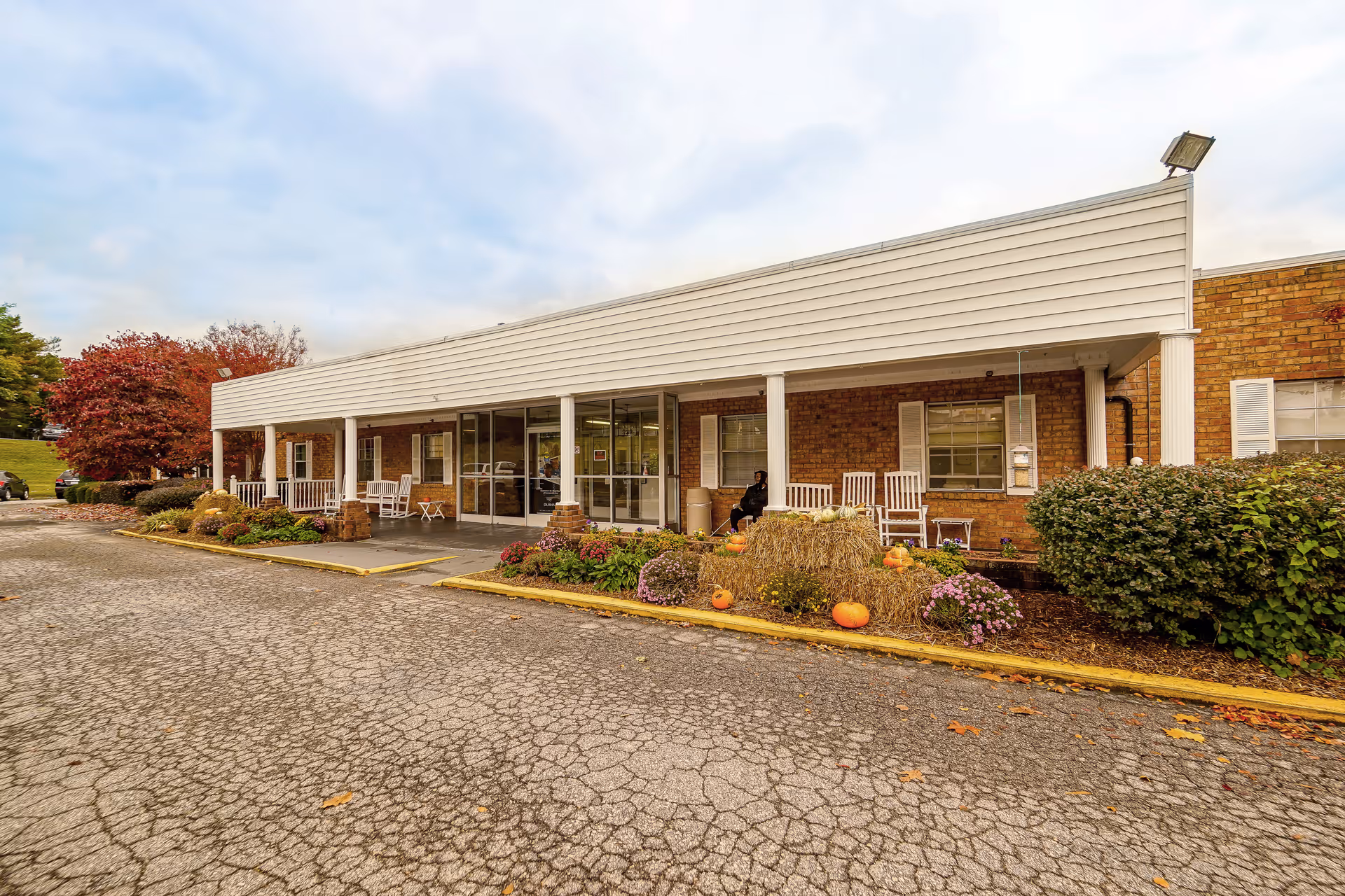 Exterior view of Durham Nursing & Rehabilitation Center showing a single-story brick building with a covered porch. The porch has white columns, rocking chairs, and seasonal decorations including hay bales and pumpkins. There are bushes and flowers along the front, and a cracked paved driveway in the foreground under a cloudy sky.