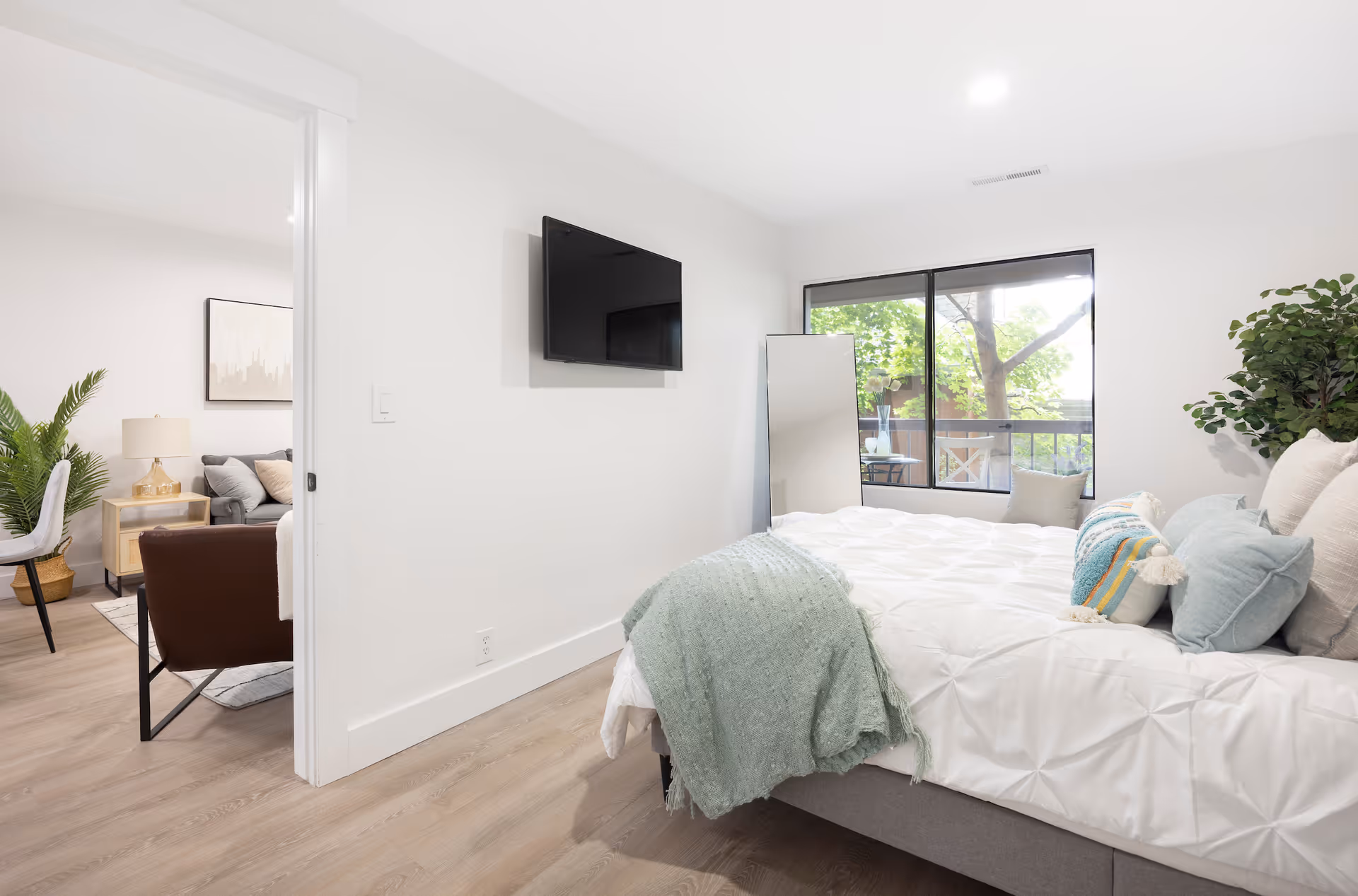 A bright bedroom with a neatly made bed featuring white bedding and multiple pillows, including a blue and striped decorative pillow. A green throw blanket is draped over the foot of the bed. A large window shows green trees outside, and a full-length mirror leans against the wall near the window. A flat-screen TV is mounted on the wall opposite the bed. The room has light wood flooring and white walls. Through an open doorway, a living area with a brown chair, a gray sofa, a side table with a lamp, and a potted plant is visible.