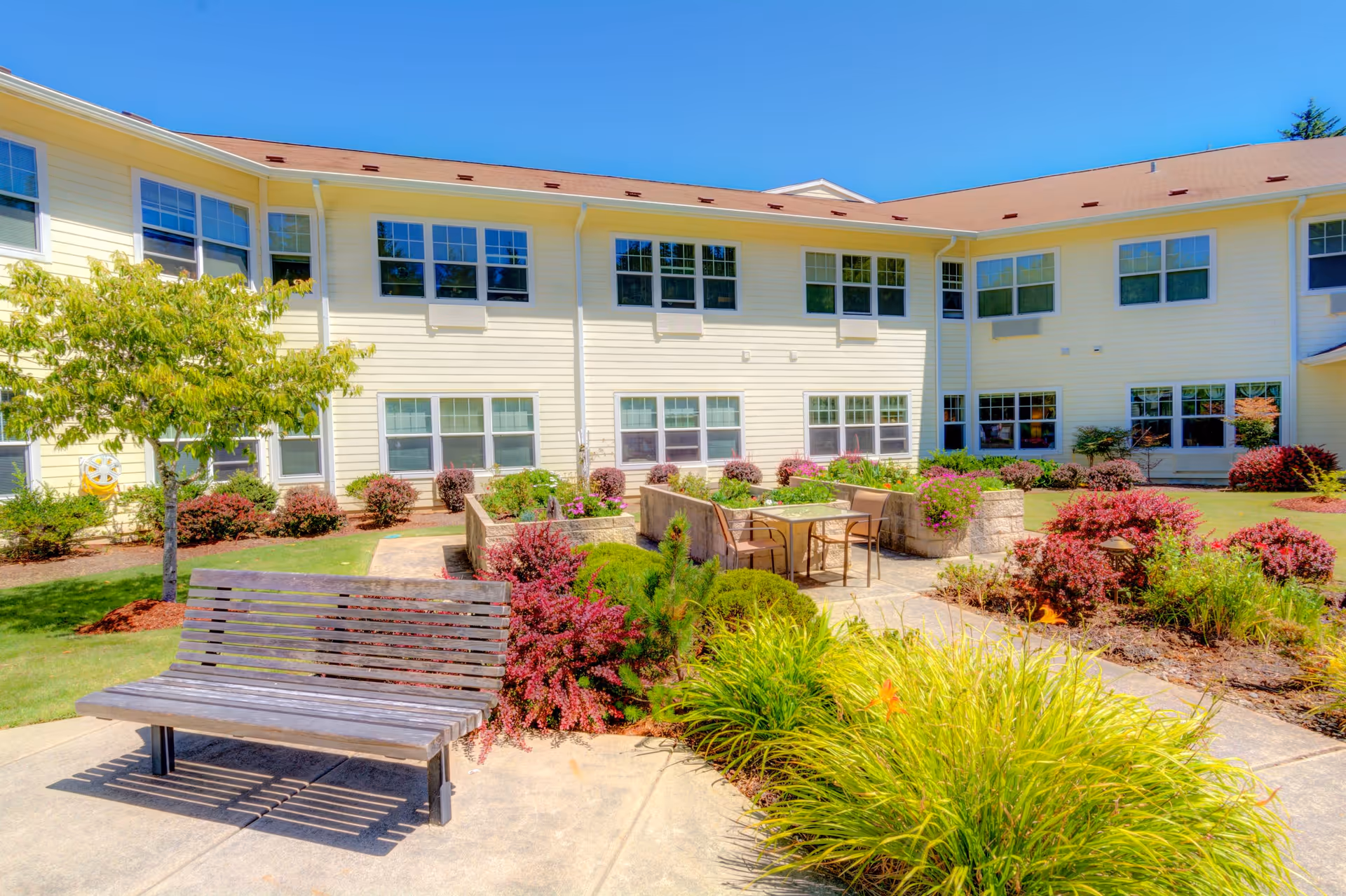Outdoor courtyard area of a senior living facility with a wooden bench, patio table with chairs, various shrubs, plants, and a tree, surrounded by a two-story yellow building under a clear blue sky.