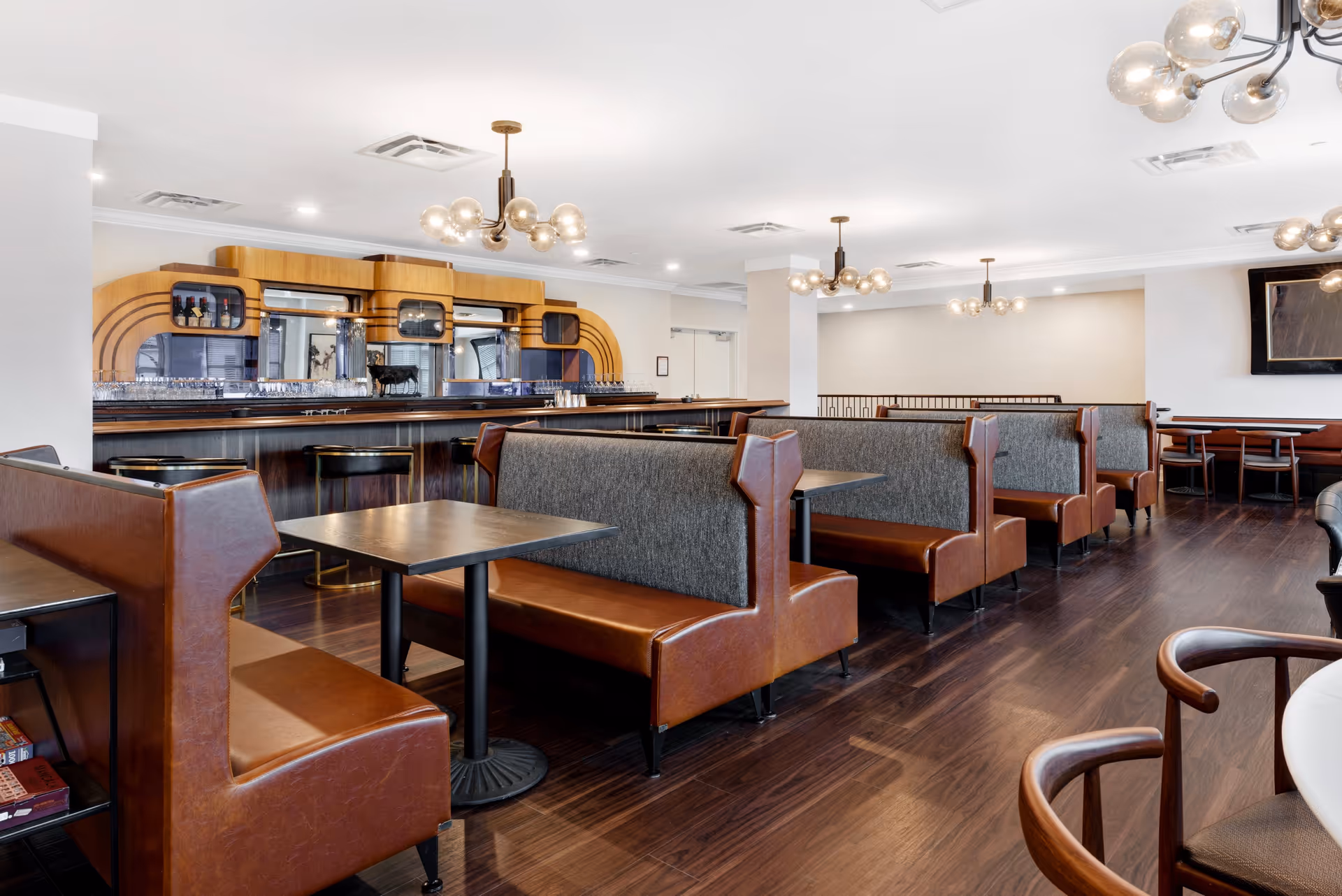 Interior view of a dining area with brown leather and gray fabric booth seating, dark wood floors, modern round chandeliers with multiple bulbs, and a wooden bar with stools in the background.
