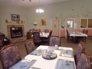 Dining room with multiple set tables and upholstered chairs, a fireplace, and wall decorations in an assisted living facility.