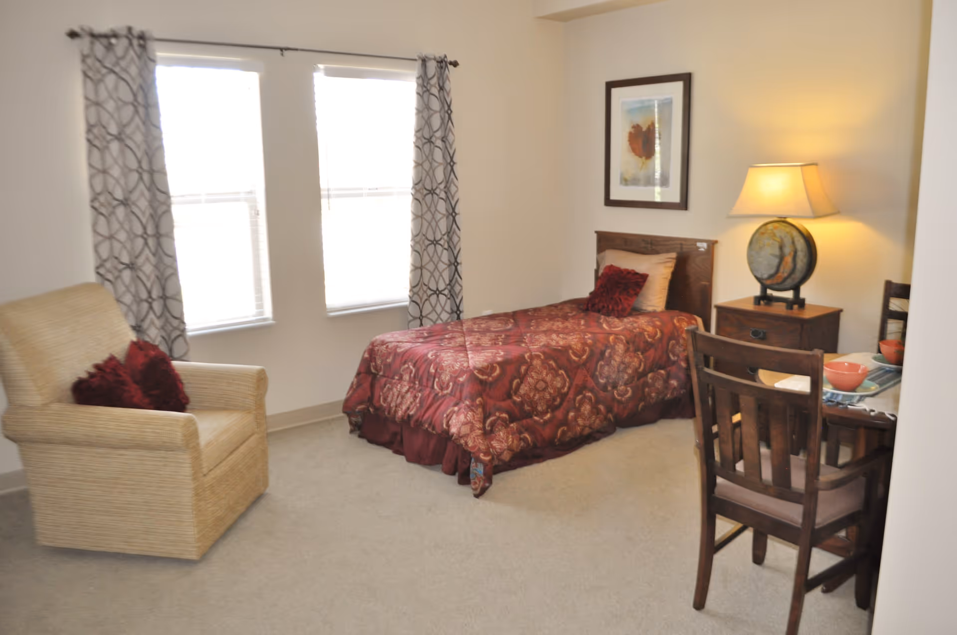 A cozy bedroom with a single bed covered in a red patterned comforter and matching pillows. Next to the bed is a wooden nightstand with a lamp and a decorative globe. There is a framed picture hanging on the wall above the bed. A beige armchair with red cushions is positioned near two windows with patterned curtains. A small wooden table with two chairs is set with bowls and napkins.