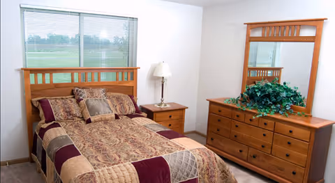 A bedroom with a wooden bed frame and a bed made with a patterned quilt and multiple pillows. Next to the bed is a wooden nightstand with a lamp. Across from the bed is a wooden dresser with a large mirror and a green plant on top. A window with blinds is visible behind the bed.