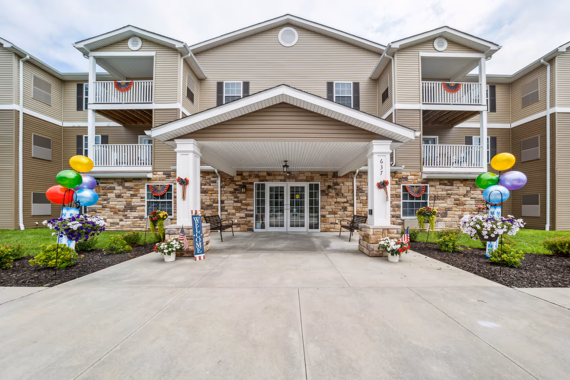 Front entrance of a multi-story retirement community building with beige siding and stone accents. The entrance has a covered porch supported by white columns, decorated with patriotic bunting and small American flags. Colorful balloons and flower arrangements flank the walkway leading to the double glass doors. Two benches are placed under the covered porch.