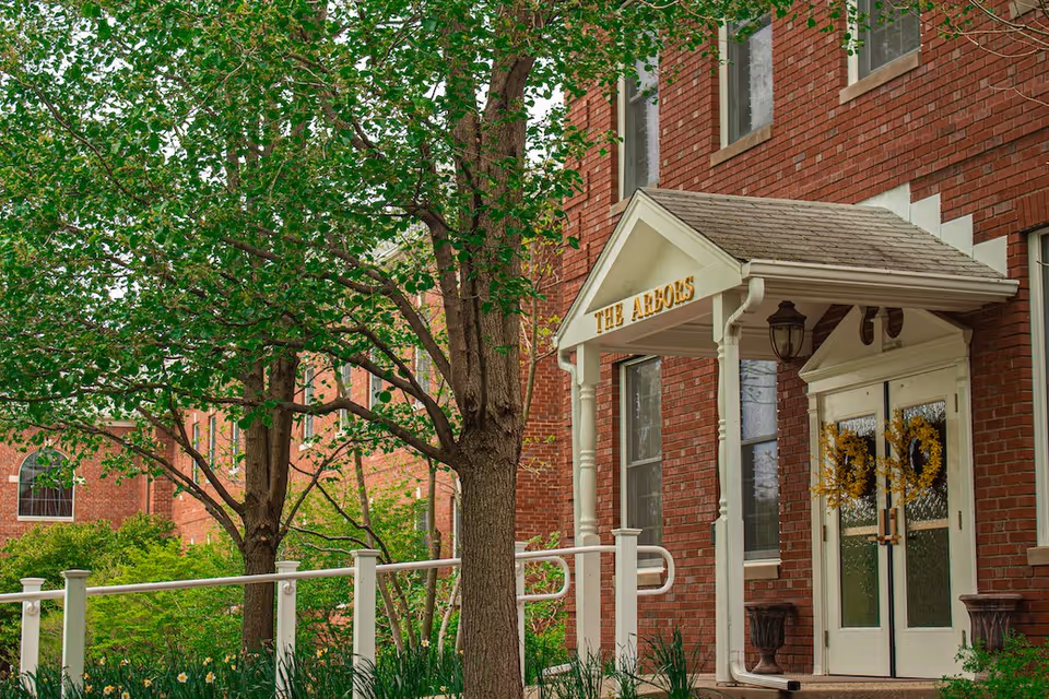 Entrance to a brick building with a white porch roof supported by columns and a sign that reads 'THE ARBORS'. The double doors have decorative wreaths, and there is a white railing along a ramp leading to the entrance. Trees and greenery surround the area.