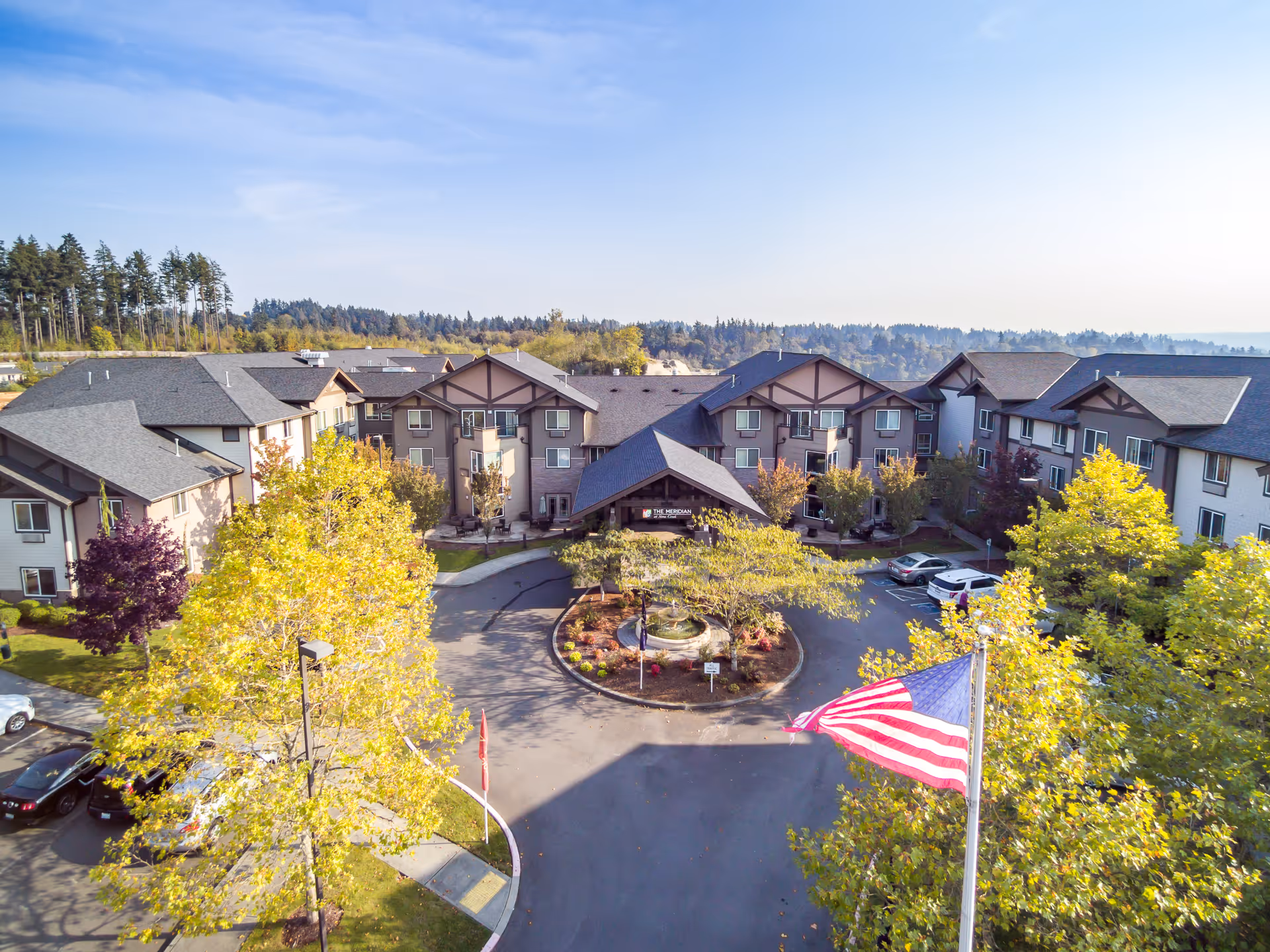 Aerial view of Meridian at Stone Creek senior living facility showing a large multi-story building with a circular driveway, landscaped garden with a fountain in the center, several parked cars, and an American flag in the foreground. Trees surround the property and a forested area is visible in the background under a clear blue sky.