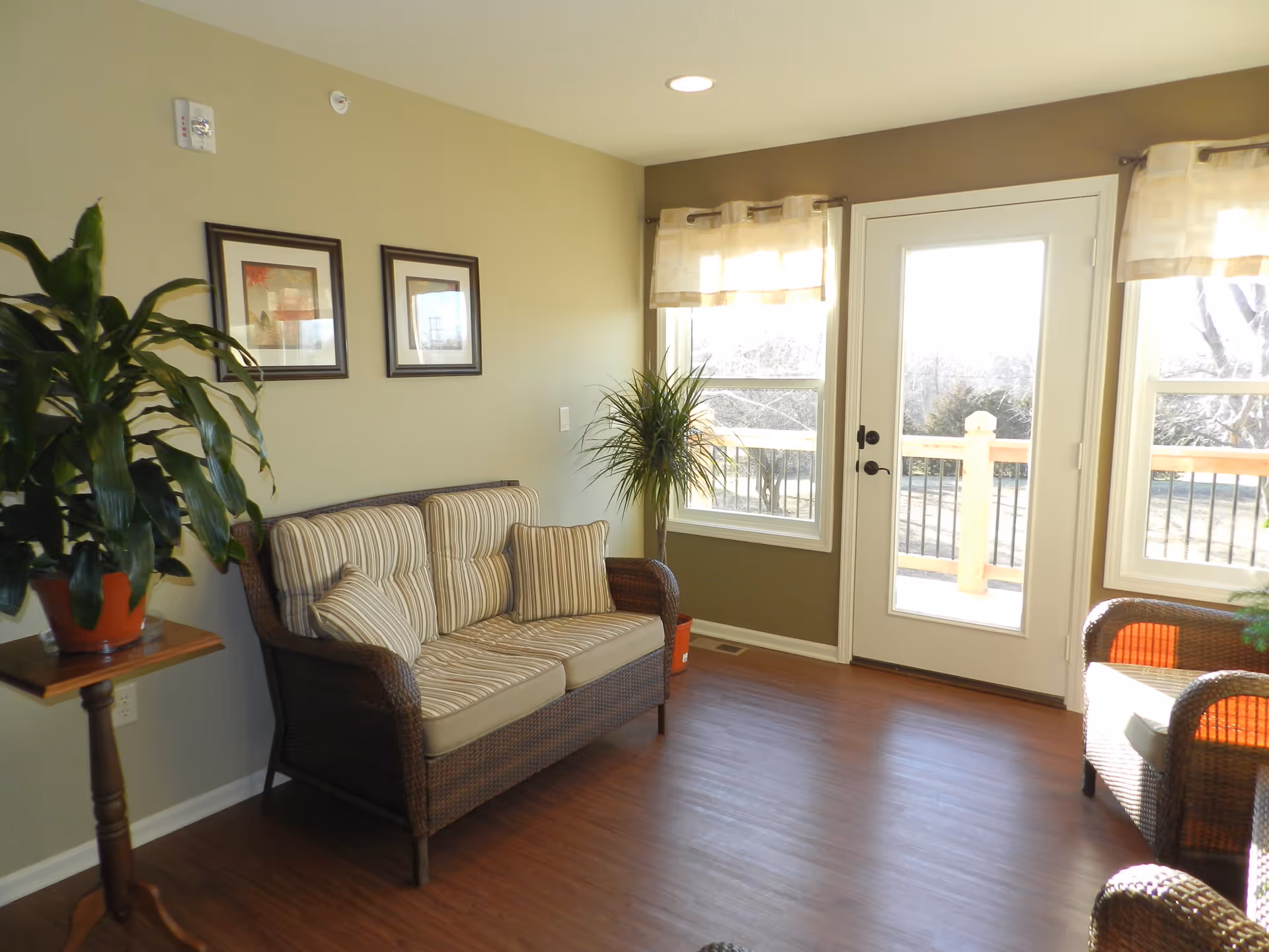 A bright living room with a wicker loveseat and armchair featuring striped cushions. There are two framed pictures on the wall, a potted plant on a wooden stand, and another plant near a glass door that leads outside. The room has wooden flooring and light-colored walls.