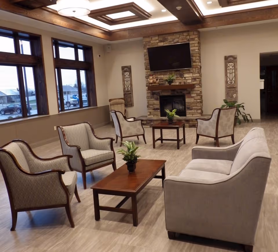 A spacious and well-lit living room area in Bay County Medical Care Facility featuring a stone fireplace with a mounted flat-screen TV above it. The room has multiple upholstered chairs and a sofa arranged around two wooden coffee tables, each with a small potted plant. Large windows with wooden frames allow natural light to enter, and the ceiling has decorative wooden beams with recessed lighting.