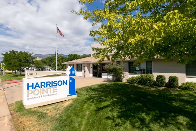 Exterior view of Harrison Pointe Healthcare and Rehabilitation building with a sign displaying the facility name and address 3430. The building is surrounded by green grass, bushes, and trees, with an American flag on a flagpole in front.