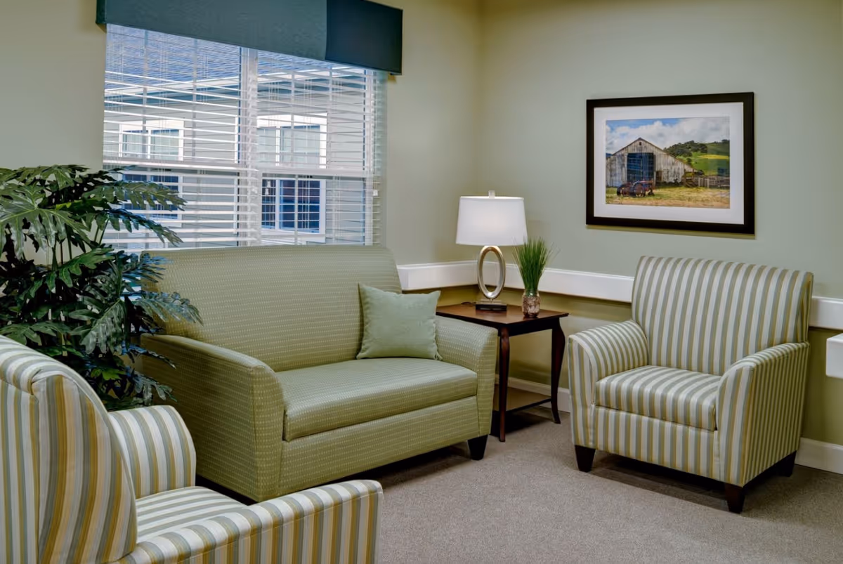 A cozy sitting area in a senior living facility with a green loveseat, two striped armchairs, a wooden side table with a modern lamp and a small plant, a large window with blinds, and a framed picture of a barn on the wall.