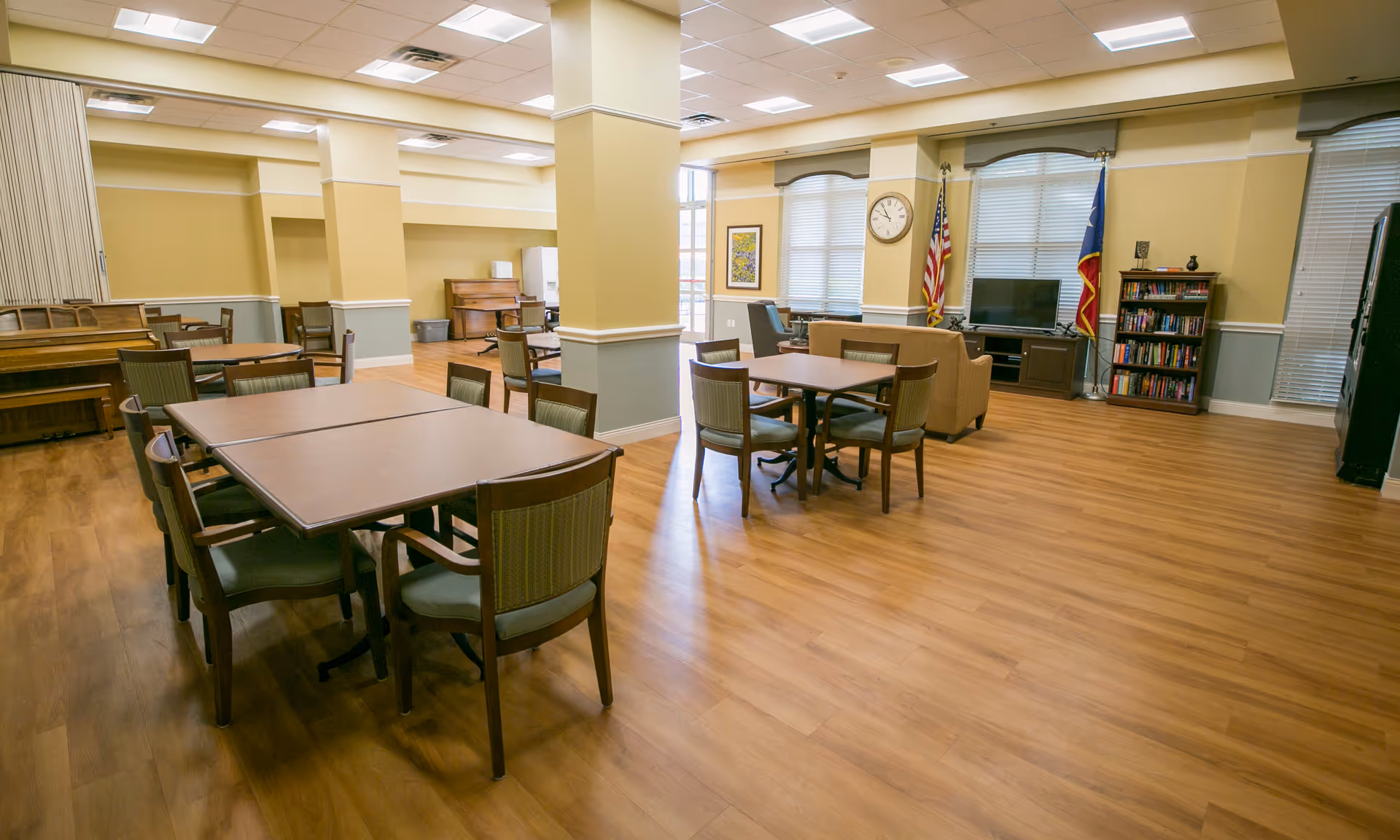A spacious common area in a senior living facility with multiple tables and chairs arranged for group seating. The room features wood flooring, beige and light blue walls, and a ceiling with recessed lighting. There is a piano against one wall, a clock, two flags, a television on a stand, and a bookshelf filled with books near large windows with blinds.