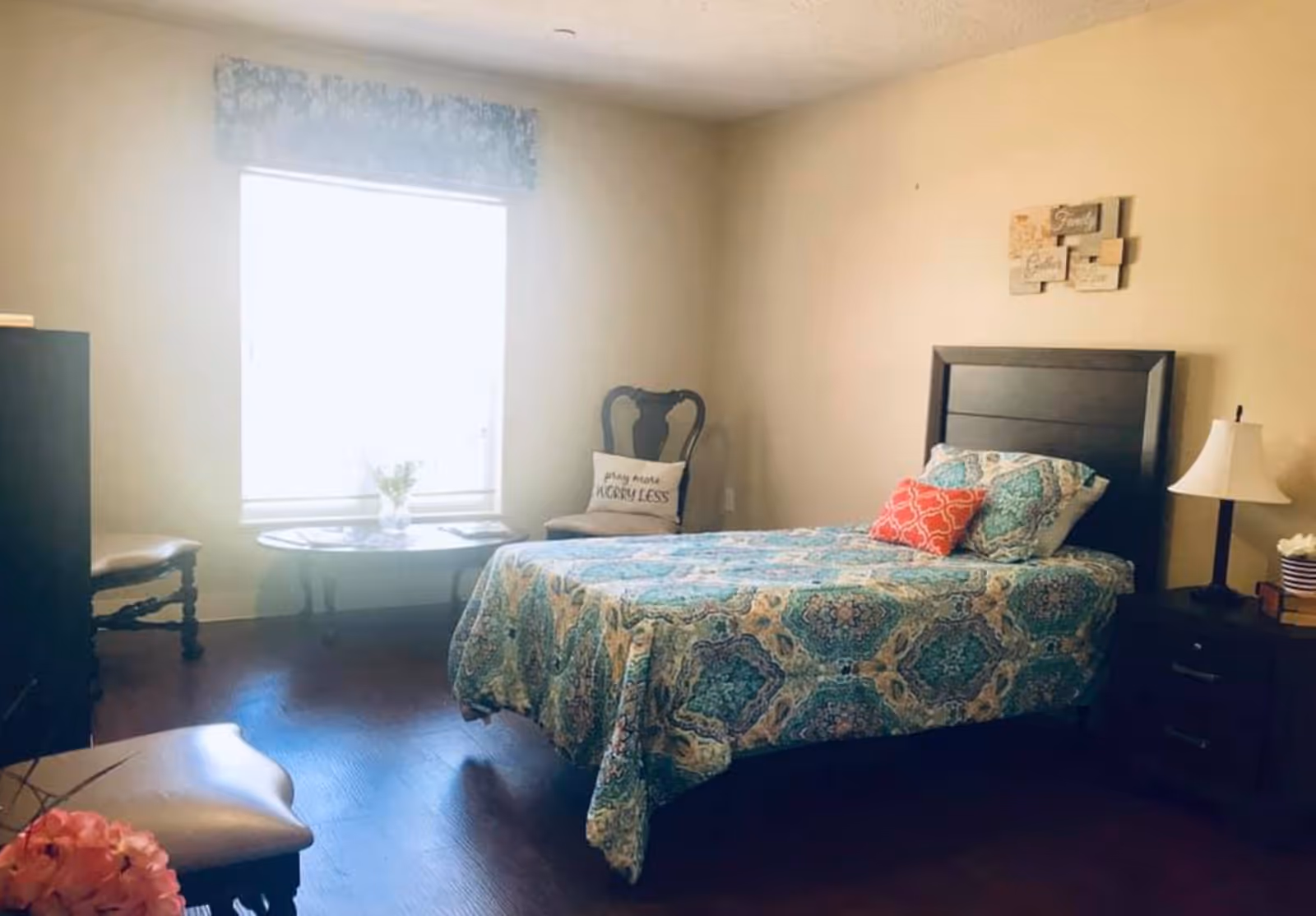 A bedroom with a single bed covered in a blue and green patterned bedspread and two pillows, one red and one matching the bedspread. There is a dark wooden headboard and a nightstand with a lamp and some books. A window with a blue valance lets in bright light. Two chairs and a small round table with a vase of flowers are near the window. The walls are beige and there is a decorative wall hanging above the bed.