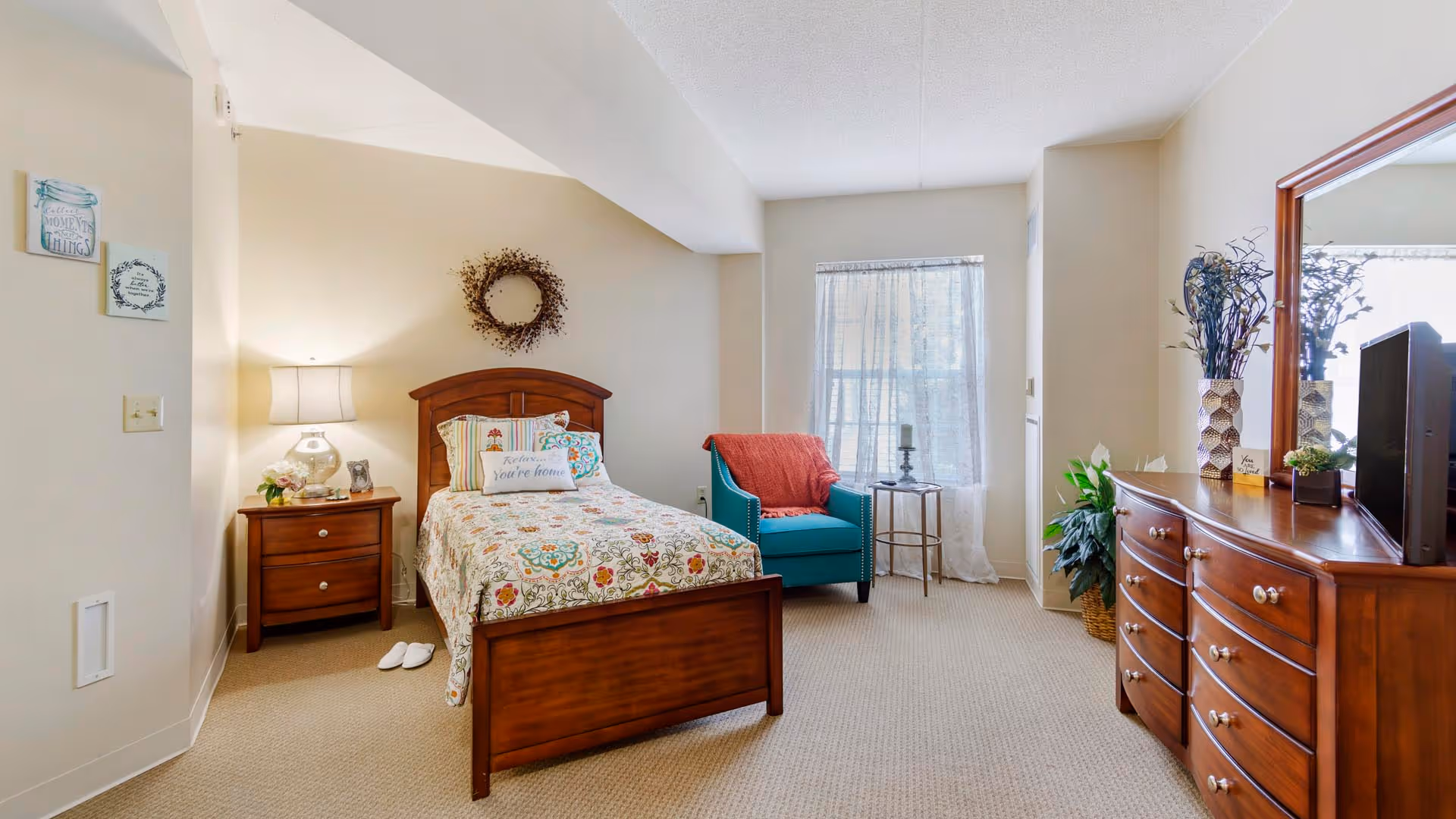 Well-lit furnished bedroom with a wooden single bed, nightstand, turquoise armchair, and wooden dresser with mirror.