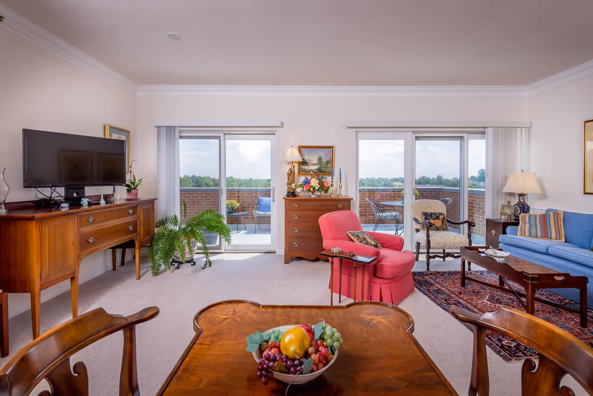 Bright living room with seating, a TV on a wooden console, sliding glass doors to a balcony, and a dining table with a bowl of fruit.