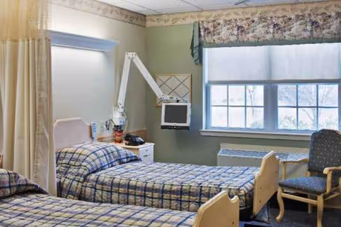 Two hospital-style beds in a shared senior living bedroom with a window and chair.