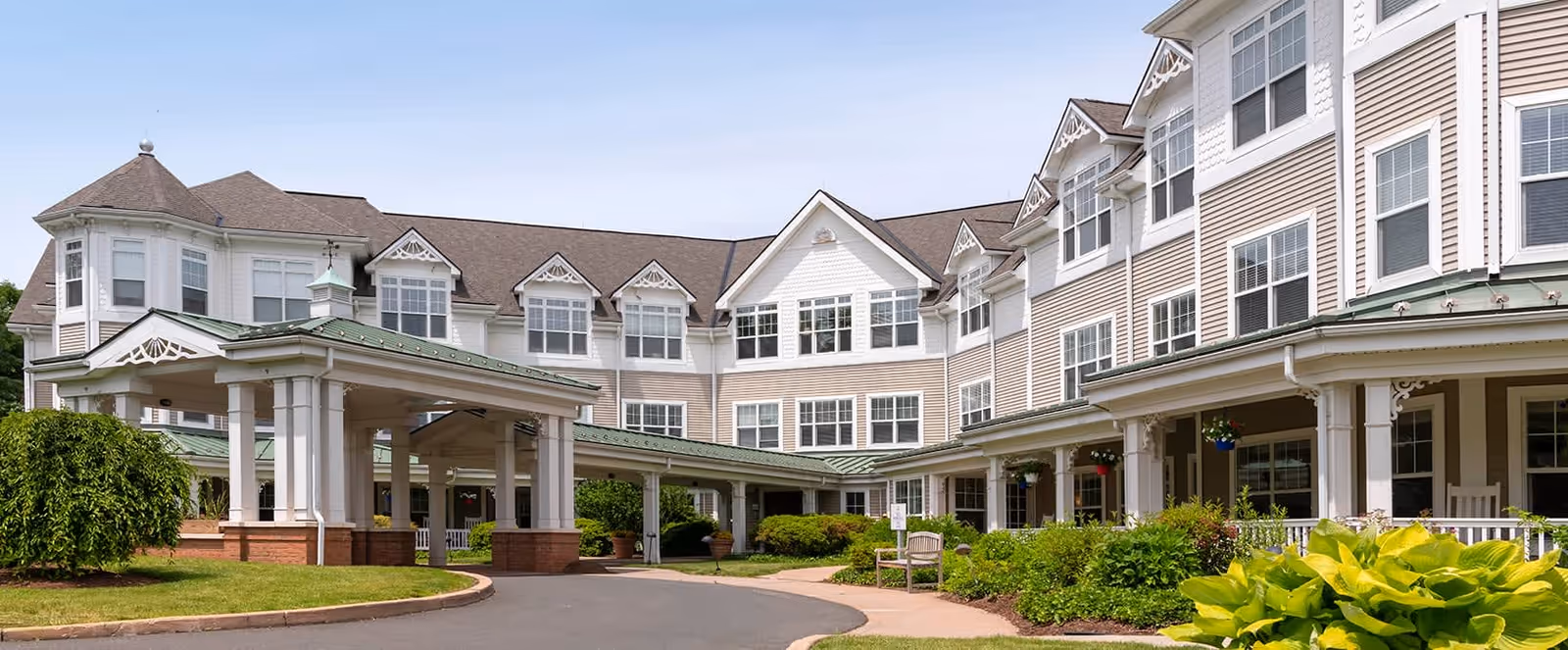 Exterior view of Brookdale West Hartford, a large multi-story senior living facility with white siding and multiple windows. The building features a covered entrance with white pillars and a green roof. There are well-maintained shrubs and greenery around the entrance and along the walkway.