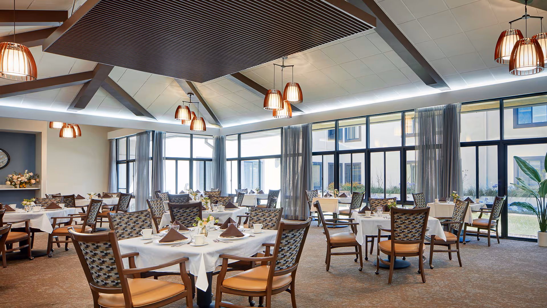 Bright, spacious dining room with multiple tables set with white tablecloths and place settings, upholstered chairs, large windows and pendant lights.