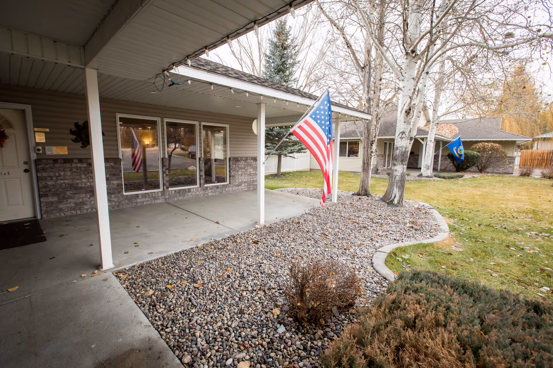 Covered porch area of a residential building with an American flag mounted on a pole. The porch has three large windows and a door to the left. The ground in front of the porch is covered with small rocks and bordered by a concrete edge. There are leafless trees and a grassy lawn surrounding the building, with another building visible in the background also displaying a flag.