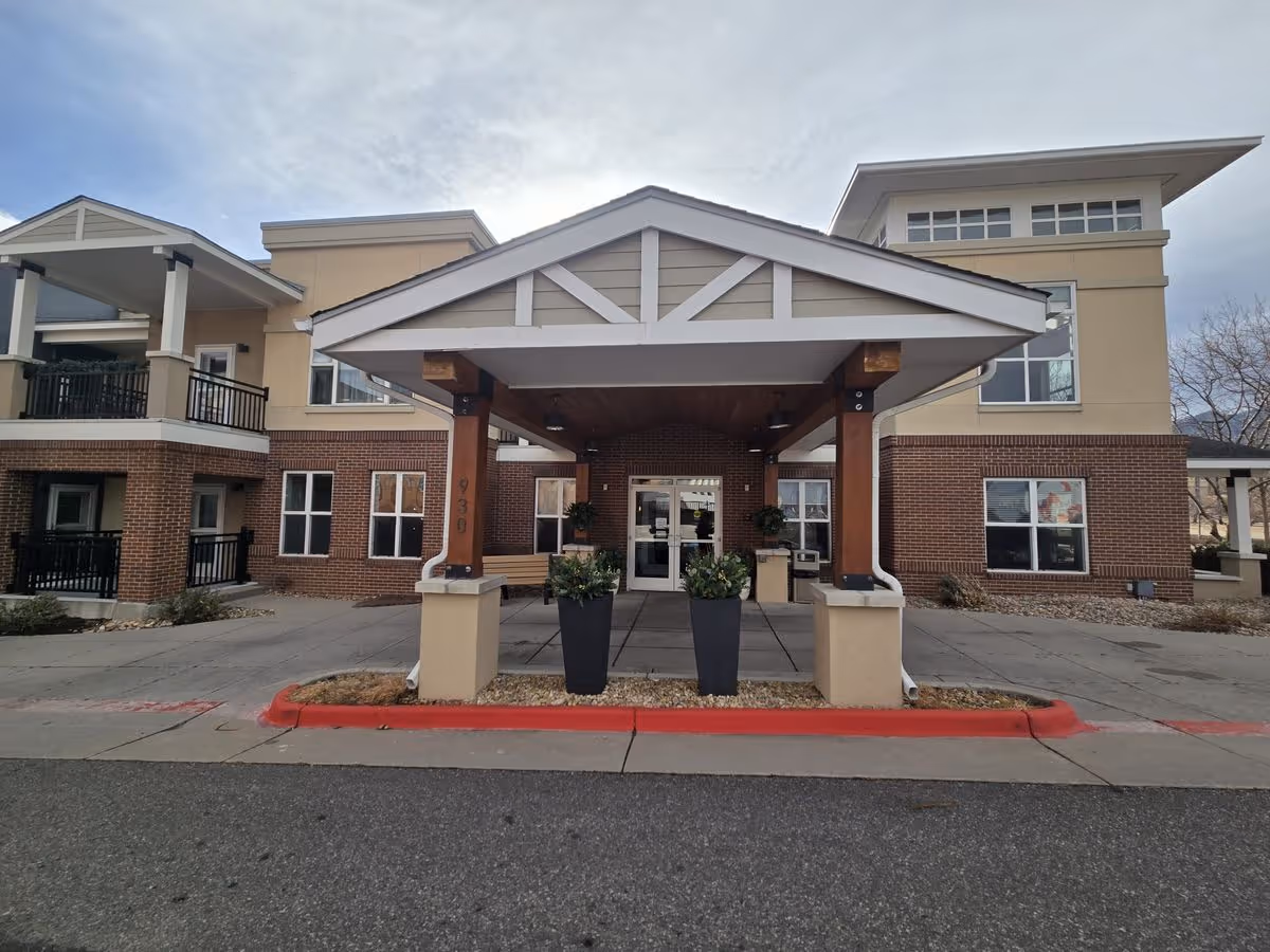 Front entrance of a senior living building with a covered porte-cochere, planters, and double glass doors.