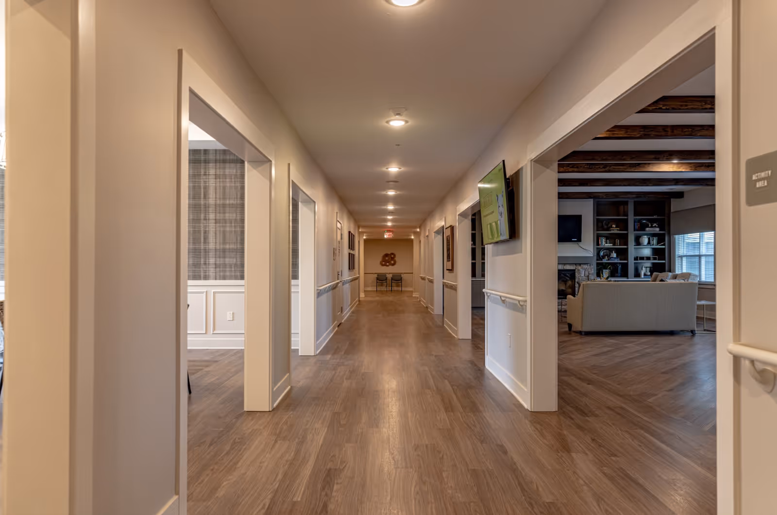 A long, well-lit hallway in a senior living facility with wood flooring and white walls. On the right side, there is an open doorway leading to a living room area with a sofa, a fireplace, and built-in shelves. On the left side, there is another open doorway showing a room with plaid wallpaper and wainscoting. Handrails run along both sides of the hallway, and ceiling lights are evenly spaced above. A television is mounted on the right wall near the living room entrance, and a sign labeled 'Activity Area' is visible on the right side.