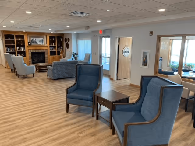 A spacious senior living common area with wooden flooring, two blue armchairs with a small wooden table between them in the foreground, a seating area with a sofa and armchairs near a wooden bookshelf and fireplace in the background, and large windows letting in natural light.