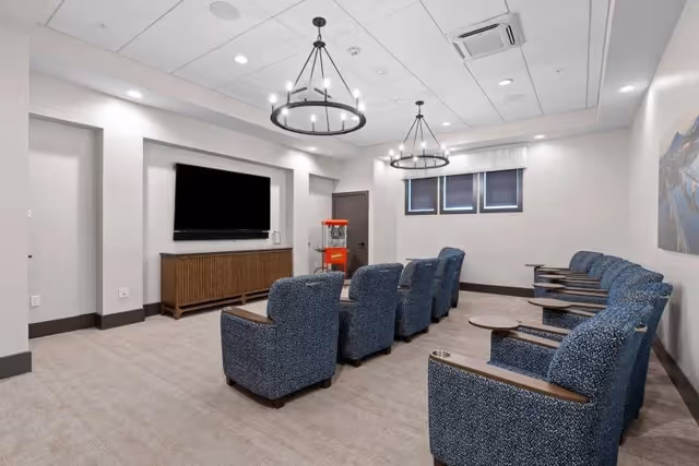 Small screening room with two rows of blue upholstered chairs facing a wall-mounted flat-screen TV and a popcorn machine.