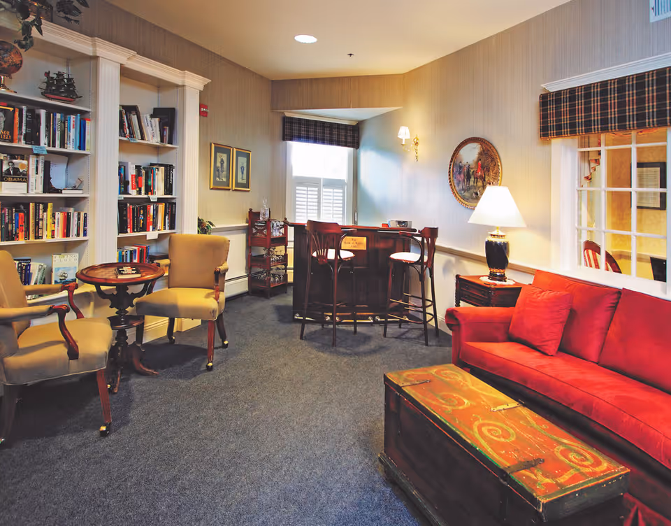 A cozy living room area featuring a red sofa with matching cushions, a wooden chest used as a coffee table, two beige armchairs around a small round wooden table, a bookshelf filled with books, a small bar with three high chairs, a table lamp on a side table, and framed artwork on the walls. The room has carpeted flooring and windows with plaid valances.