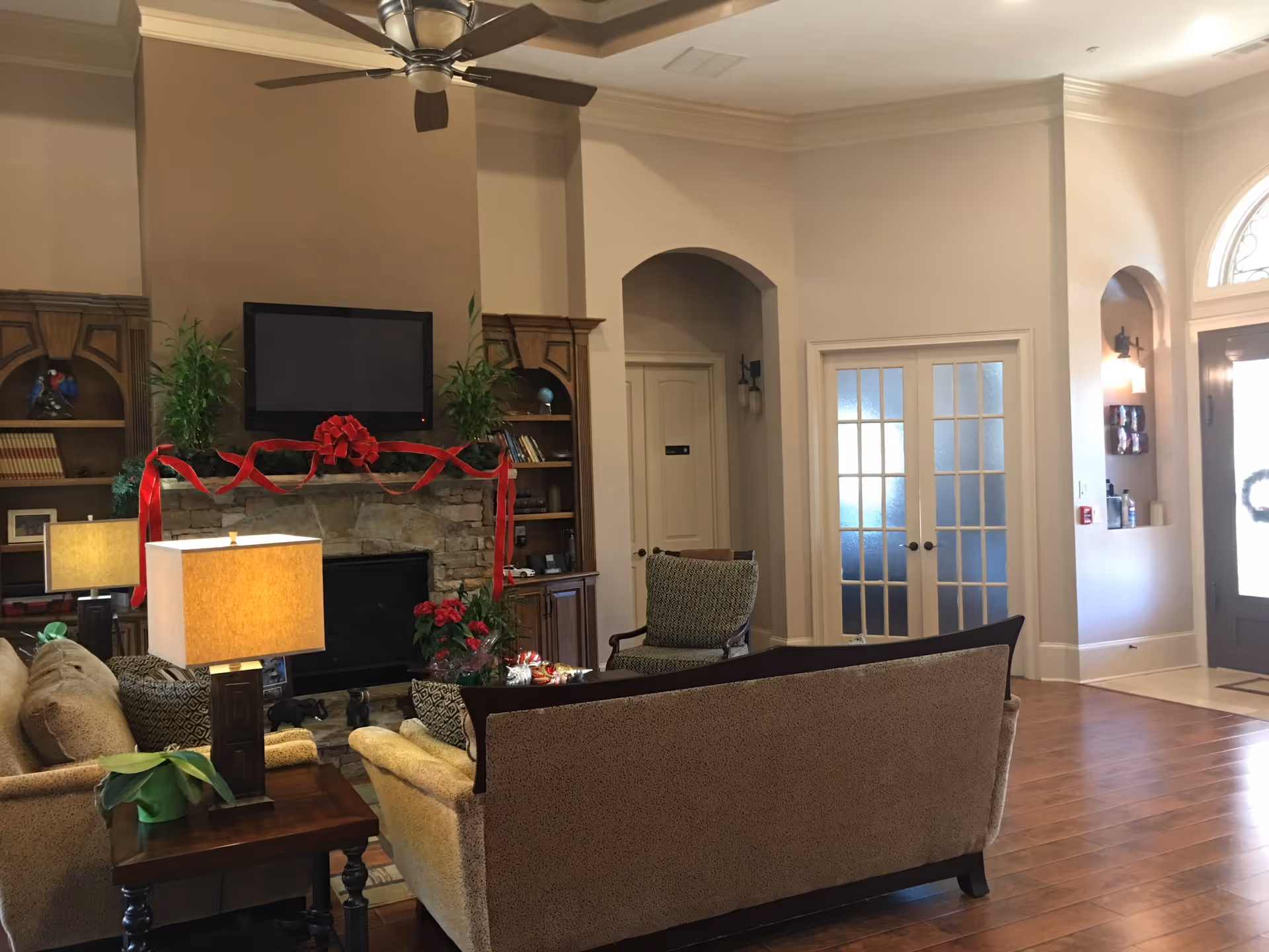 A cozy living room area in a senior community featuring a stone fireplace decorated with a red ribbon and bow, a flat-screen TV mounted above the fireplace, wooden built-in shelves with books and plants, beige upholstered sofas and chairs, a wooden side table with a lamp and a potted plant, hardwood flooring, and a ceiling fan. French doors and an entrance door with a wreath are visible in the background.