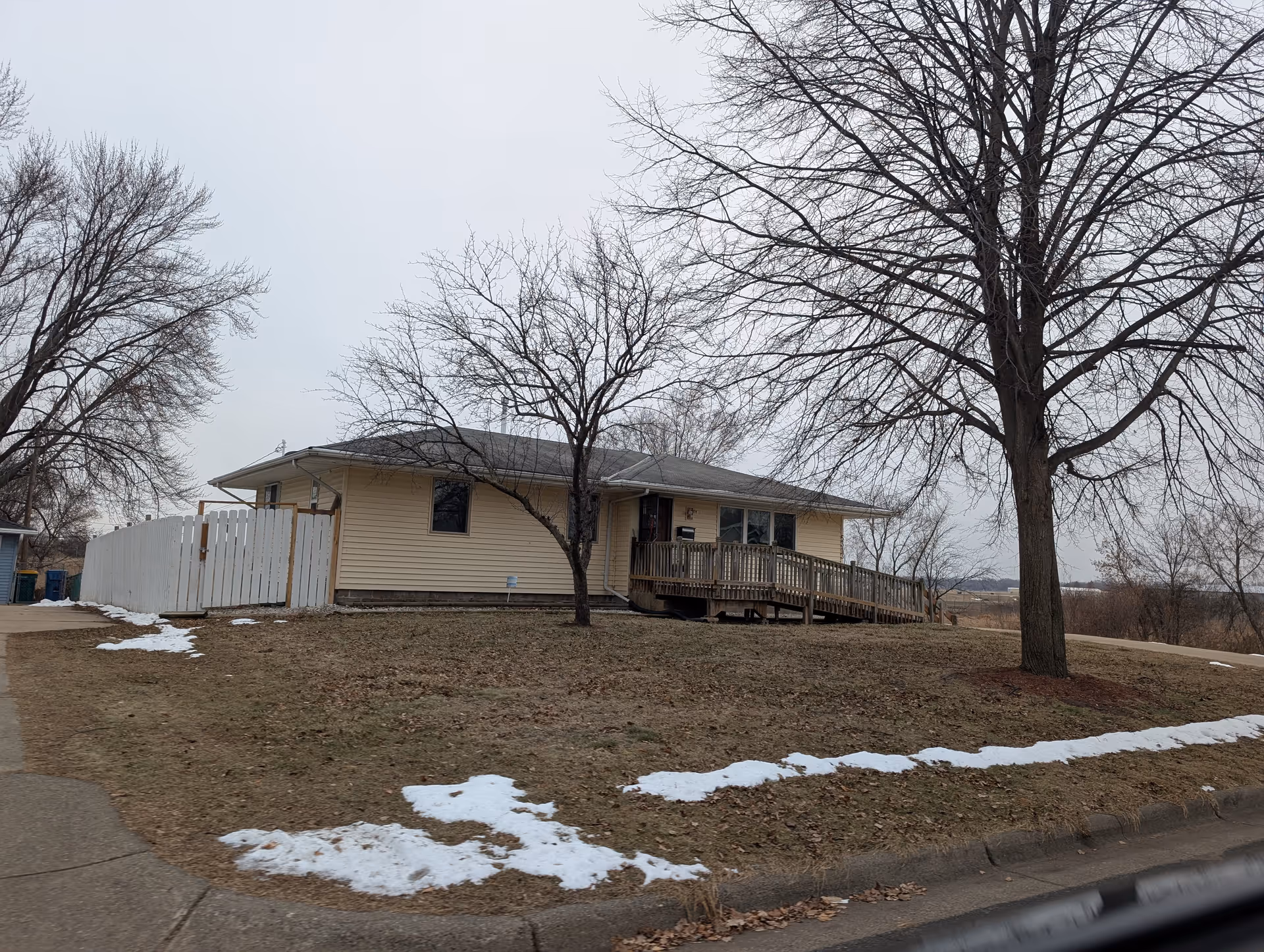 Single-story beige building with a wheelchair ramp at the entrance, surrounded by leafless trees and patches of snow on the ground under an overcast sky.