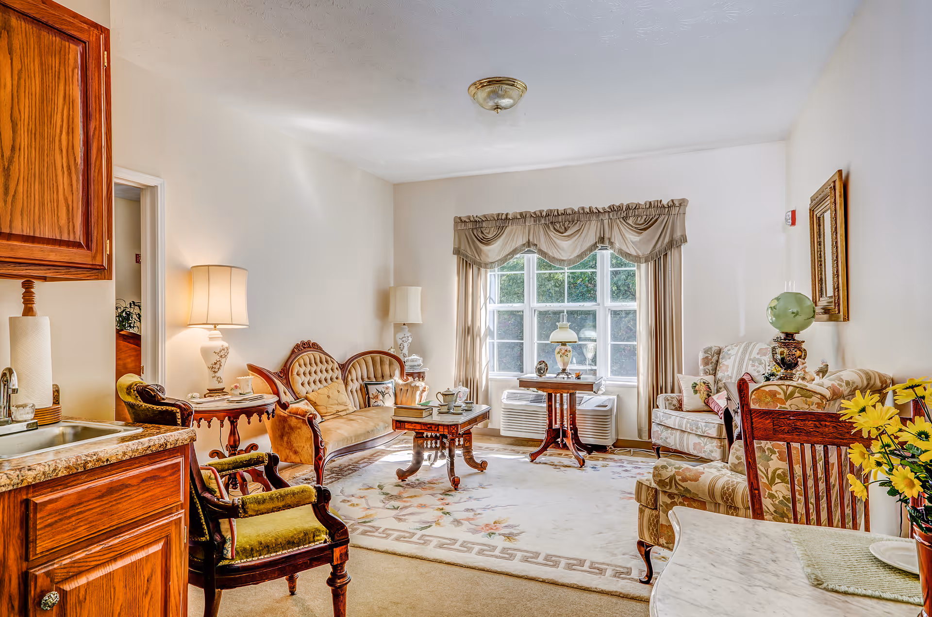 A bright and cozy living room in a senior living facility featuring vintage-style furniture including a tufted sofa, armchairs, a wooden coffee table, and side tables with lamps. A large window with draped curtains lets in natural light, and a patterned area rug covers the floor. The room also includes a small kitchenette area with wooden cabinets and a sink.