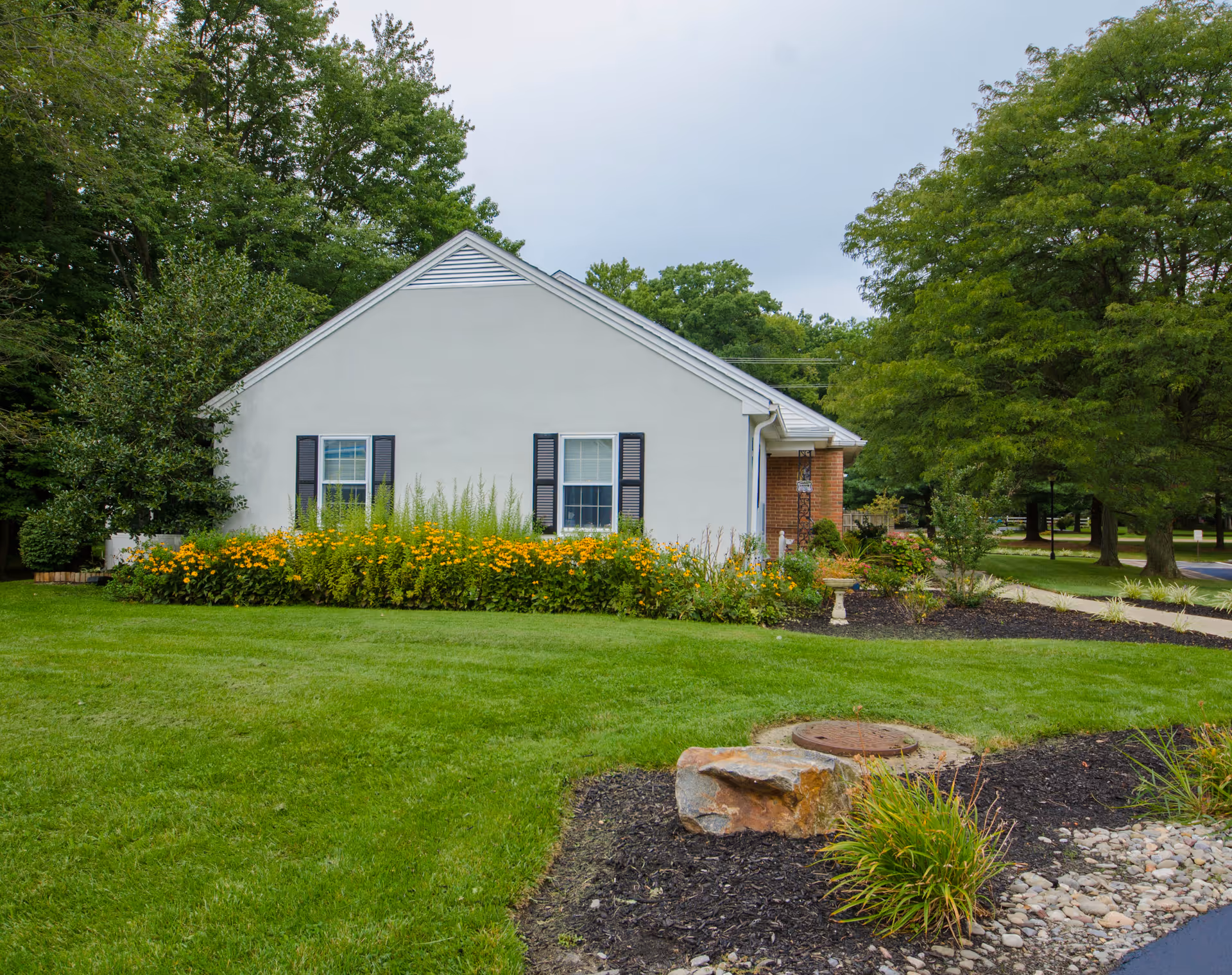 Exterior view of a single-story building with white walls and black window shutters, surrounded by green grass, trees, and a garden with yellow flowers and shrubs.