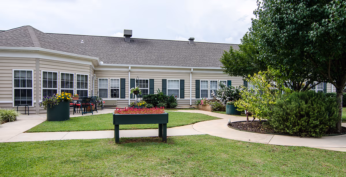 Outdoor view of a senior living facility with beige siding and multiple windows with dark green shutters. There is a well-maintained lawn with a raised flower bed in the center, several potted plants with colorful flowers, a curved concrete walkway, and trees and shrubs on the right side.