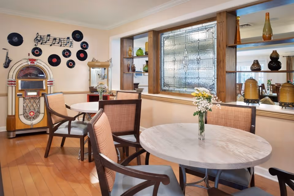 A cozy dining area with round tables and cushioned chairs. Each table has a small vase with white flowers. The room features a decorative jukebox and a wall adorned with vinyl records and musical notes. There is a large stained glass window with wooden shelving displaying various vases and decorative items.