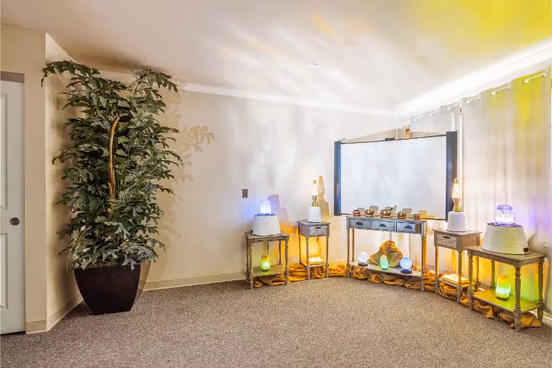A softly lit room with a large potted plant on the left, three small tables with glowing decorative lights, and a larger table with a mirror and small cups arranged on top. The room has beige walls, carpeted floor, and sheer curtains on the right side.