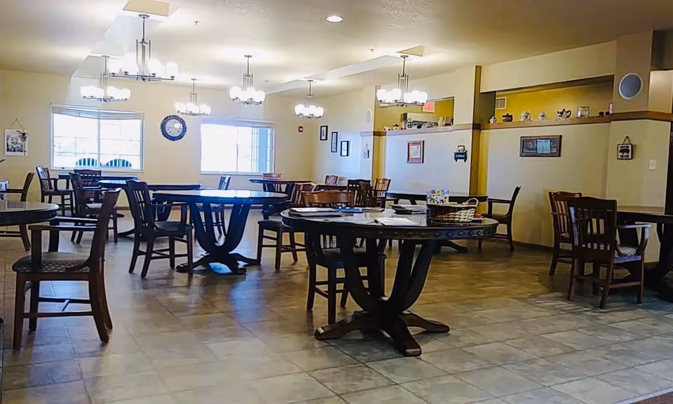 A spacious dining room with multiple round and rectangular wooden tables and chairs arranged neatly. The room has large windows letting in natural light, beige walls with framed pictures and a clock, and several ceiling light fixtures providing illumination.